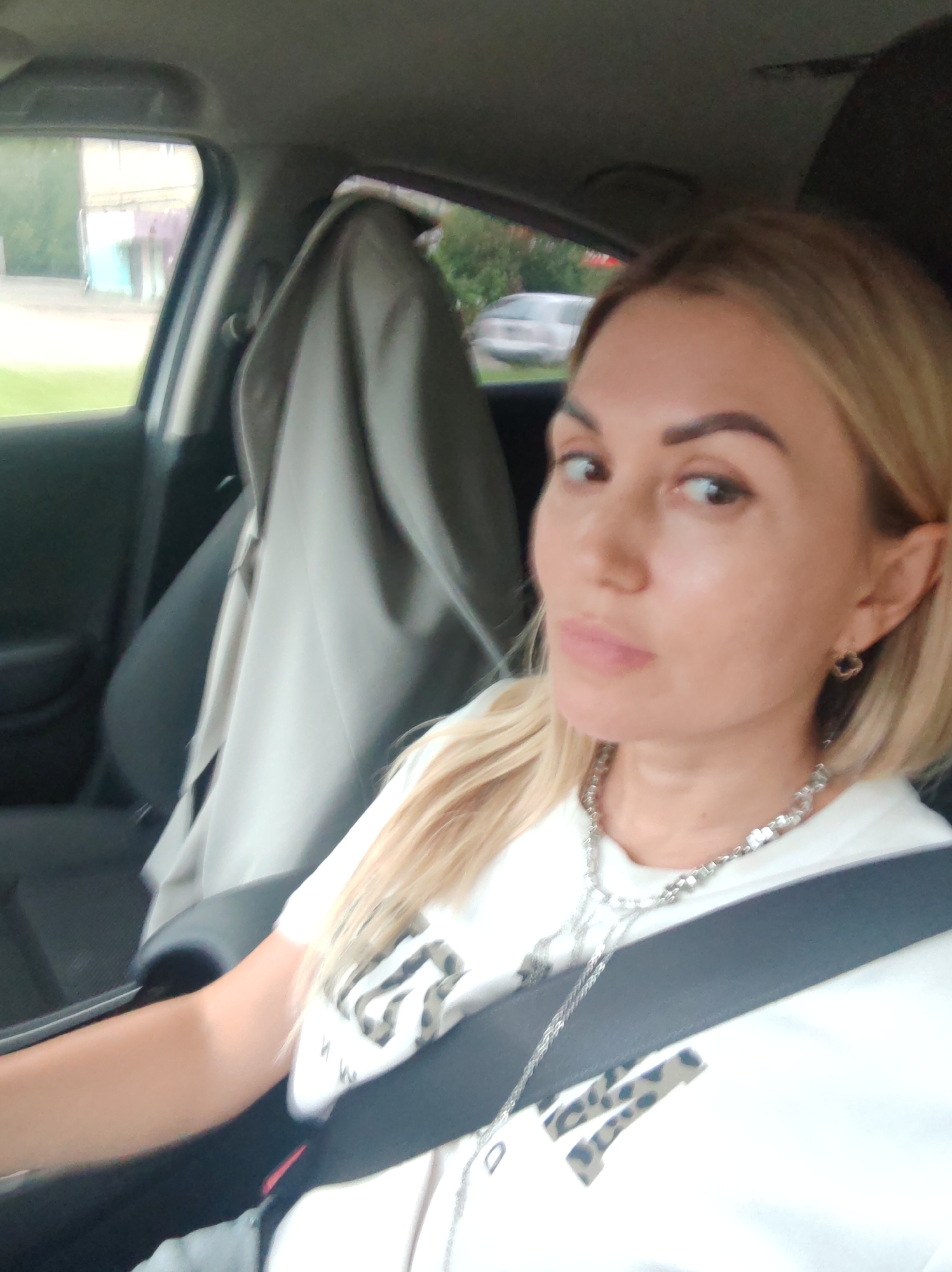 The image shows a woman in the driver's seat of a car. She has blonde hair and is wearing a white t-shirt with a leopard print design. She is also wearing a silver necklace and hoop earrings. The woman is looking at the camera with a neutral expression. The car appears to be parked, with the window down and a beige jacket or blanket draped over the passenger seat. Outside the car, there are trees and buildings visible, suggesting an urban or suburban setting. The lighting suggests it is daytime, possibly overcast. The image is taken from a selfie perspective.