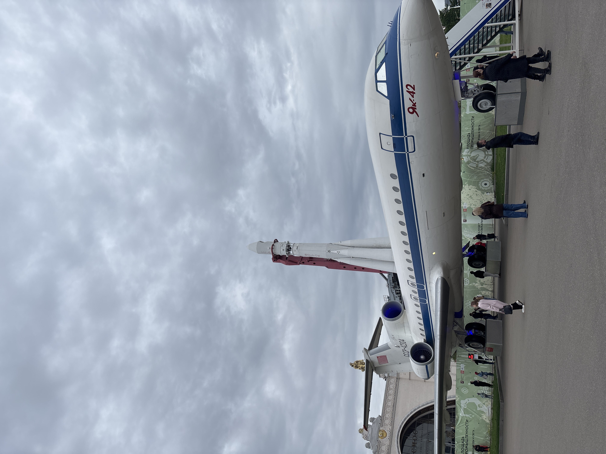 The image shows a Yak-42 aircraft on display, next to a rocket, at VDNH, an exhibition center in Moscow, Russia. People are standing near the plane. The sky is cloudy. The plane has the registration number "COOP-42304" on it. The rocket is labeled "BOCTOK." Signage nearby contains Russian text.