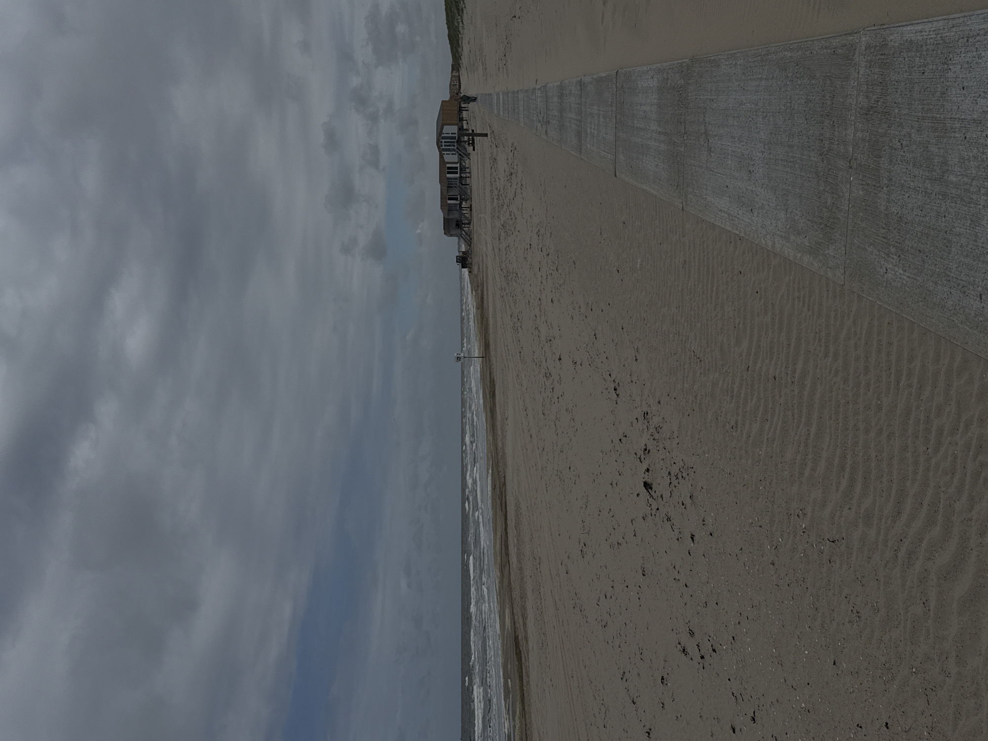 The image displays a wide, expansive beach scene along a coastline under a heavily overcast sky. The horizon line is visible on the left side, where the grey sea meets the sandy shore with small, breaking waves and whitecaps. A vast stretch of light brown sand, showing textured ripples and scattered debris, occupies the central and right portions of the frame.
Running parallel to the beach on the far right is a long, grey concrete path or promenade, constructed from rectangular slabs. Along this path, in the middle distance, several elevated beach houses or bungalows are visible. These structures feature wooden exteriors, dark roofs, and stairs leading up to their entrances, situated on the edge of the sand. Faint hints of green dune vegetation can be seen behind these buildings.
The sky, filling the upper left section, is dominated by thick, grey clouds, though some lighter areas suggest variations in cloud cover. No people are visible, and the beach appears deserted, conveying a quiet atmosphere. A slender pole or marker stands in the sand closer to the water. The scene depicts a calm, spacious coastal landscape, likely daytime given the overall light, but with subdued weather conditions. There is no visible text in the image.