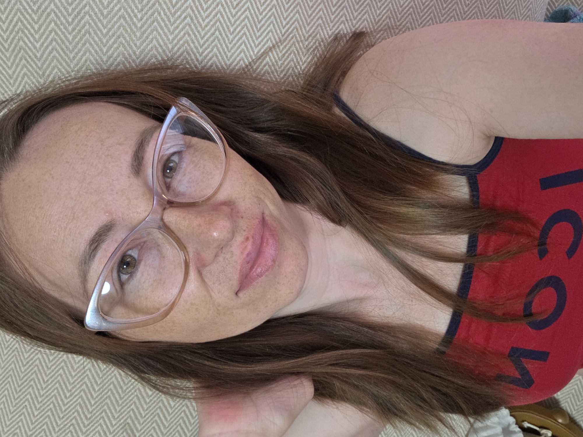 A woman with auburn hair and freckles is lying down, possibly on a beige patterned couch or bedspread. She is wearing glasses with a light-colored frame and a red tank top with black trim. A portion of the writing on the tank top is visible, appearing to be "COM." Her skin is fair with a pinkish hue, and she is looking at the camera. There is no visible indication of the location being the city of Chaikovsky, Russia.