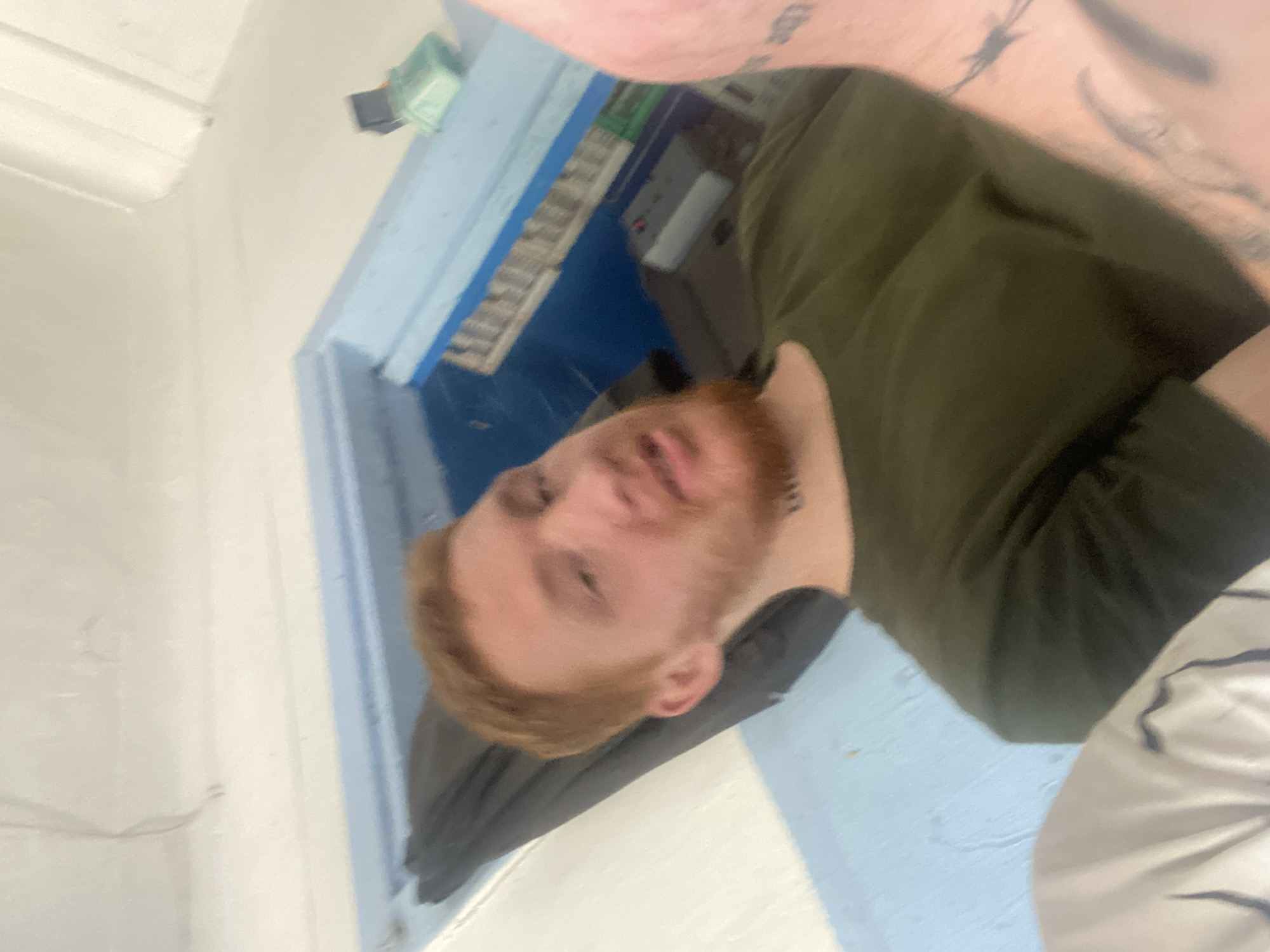 The image is a selfie taken by a person with red hair and a beard, wearing a green t-shirt. The person is lying down, with their head and upper body visible. The background appears to be an indoor space with white walls and blue accents. There is a dark fabric object behind the person, and some electronic equipment is visible in the background, including a white box with a red light and a green object on a blue ledge. The person has a tattoo on their neck and arm. The overall lighting is bright, suggesting daytime.