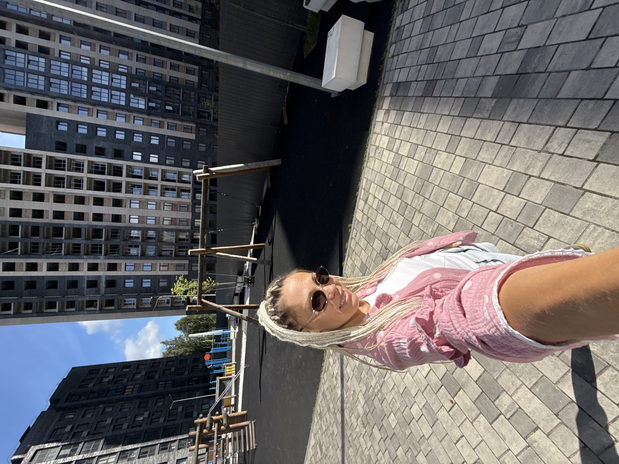 A woman with blonde braided hair and sunglasses takes a selfie outdoors on a paved walkway. She is wearing a pink patterned shirt and a white t-shirt. Behind her is a children's playground with swings and climbing structures, and in the background are large, modern apartment buildings, some of which appear to be under construction. The sky is blue with some clouds, and the sun is casting shadows. The location is identified as Rusanovo, Russia.