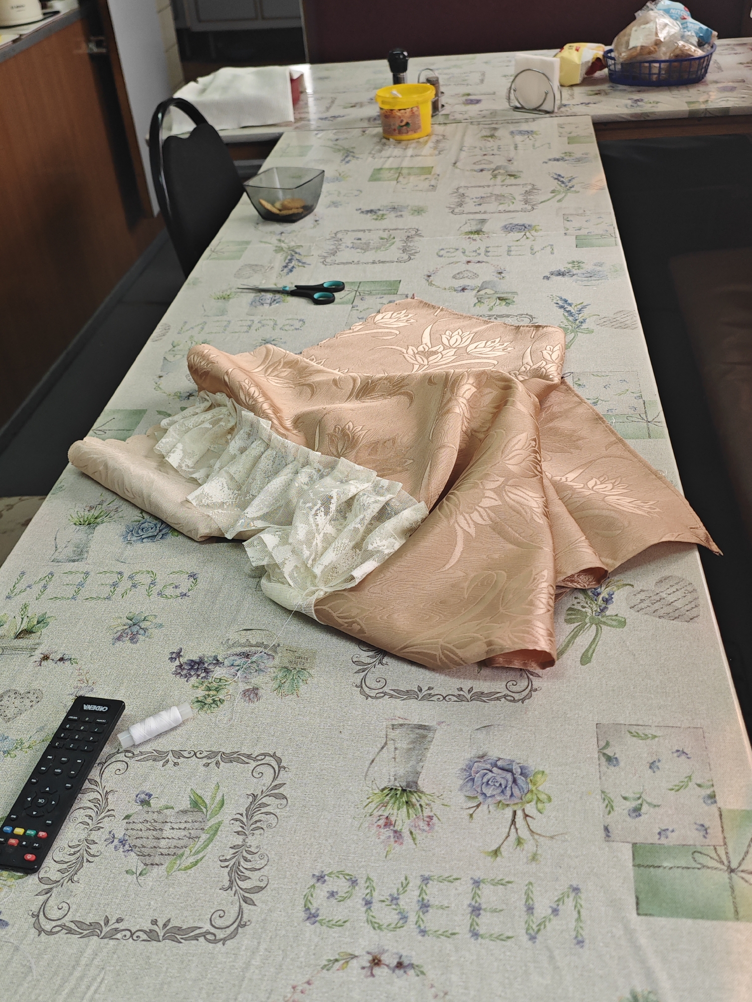 A tablecloth with a floral and script pattern is draped over a table. On the tablecloth, there are several items, including a remote control, a spool of white thread, a pair of scissors, a small bowl with biscuits, and a folded piece of patterned fabric. The fabric is a peachy-pink color with a raised floral design, and a section of white lace trim is visible. The background shows a chair and part of a kitchen or dining area. This scene appears to be a domestic setting.
