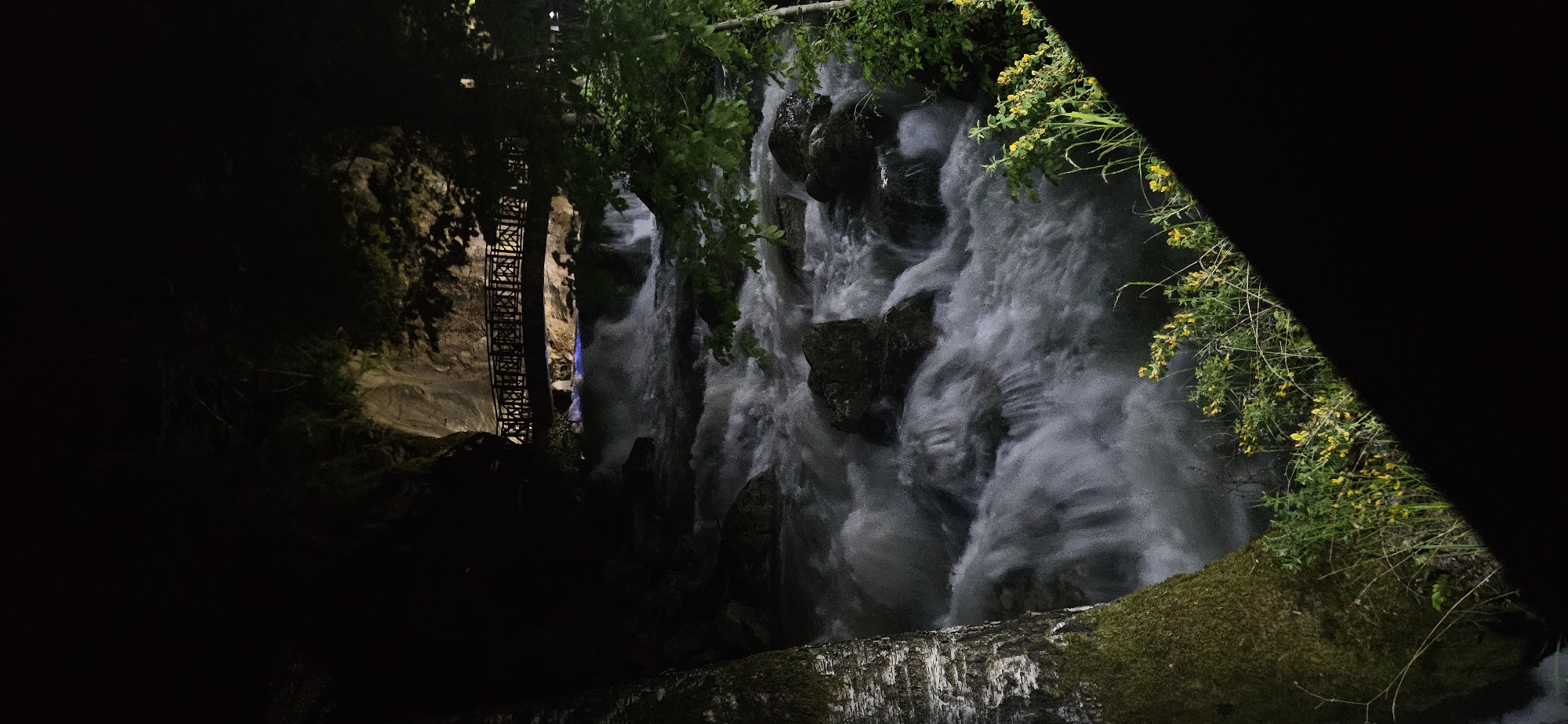 The image shows a waterfall at night in Antalya, Turkey. A metal staircase is visible on the left side of the waterfall. The water is flowing rapidly over rocks and the surrounding area is partially obscured by darkness and foliage. There are some flowers visible in the foreground.