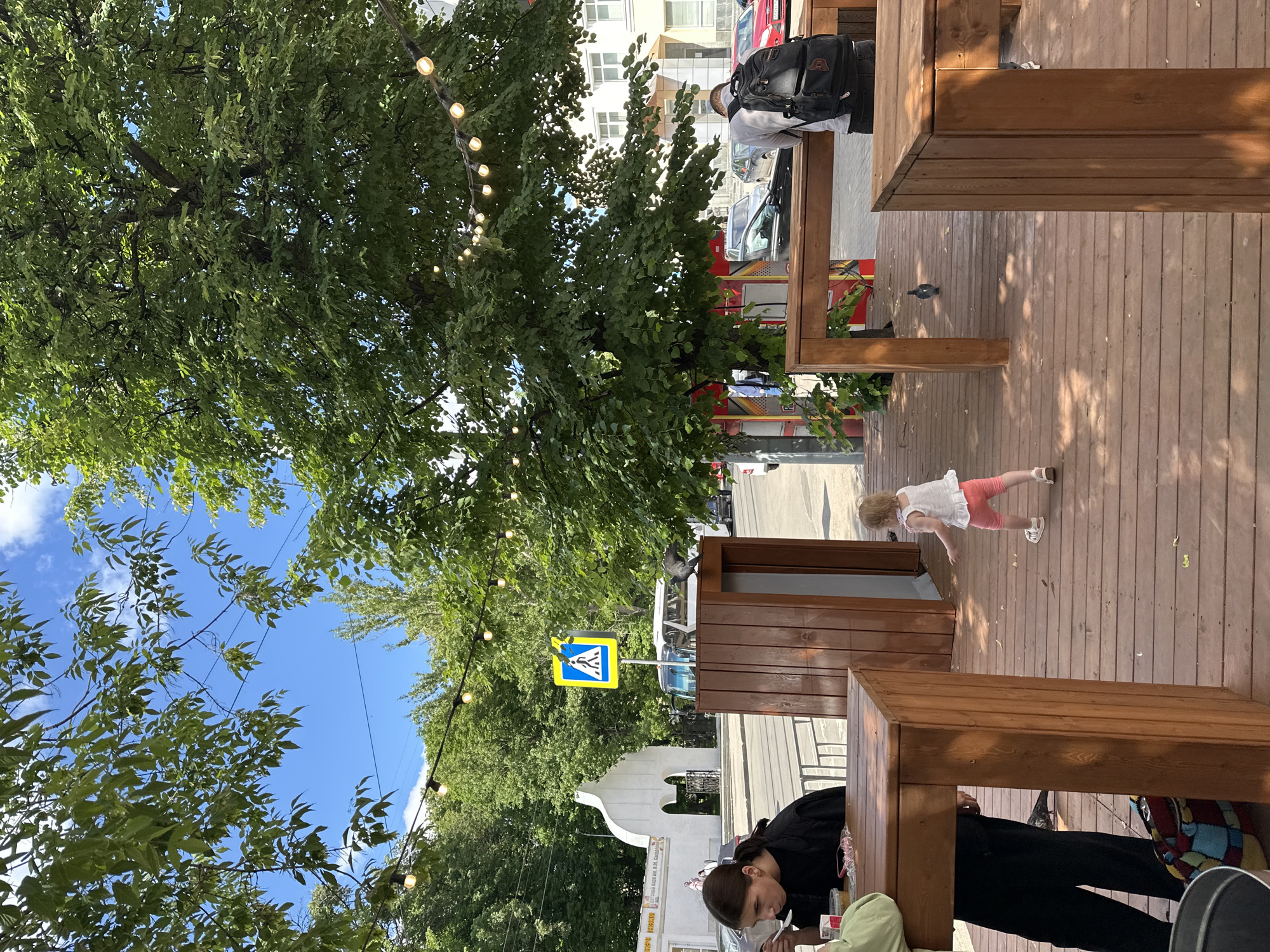 The image captures a lively outdoor scene at what appears to be a cafe or seating area in Nizhny Novgorod, Russia, on a bright, sunny day. The perspective looks up through a dense canopy of lush green trees, under which string lights are hung.
In the foreground, a young woman with dark hair, dressed in black, is seated at a wooden table on the left, looking down at a bowl she is eating from with a spoon. A light green object is on the table beside her. The seating area is constructed from wooden decking and features sturdy wooden tables and benches.
In the mid-ground, a small child, a girl with blonde hair wearing a white top and coral shorts, is actively engaged on the wooden deck, leaning forward on her hands and one foot, seemingly playing. A pigeon is also visible on the decking further back.
Beyond the seating area and trees, a street scene unfolds. Cars are visible, and in the distance, modern buildings stand alongside an older, white building with an arched entrance. Partial Cyrillic text can be seen on a sign on this white building, including "Курица" (Chicken). A prominent blue square pedestrian crossing sign with a white triangle and black pedestrian symbol is visible near the street. A man wearing a light shirt and dark backpack is seen standing or walking near the street in the background.
The sky is clear blue with scattered white clouds, suggesting pleasant weather. Shadows from the trees are dappled across the wooden deck. The string lights, while visible, are not illuminated, indicating it's daytime. The overall impression is one of a relaxed urban outdoor space.