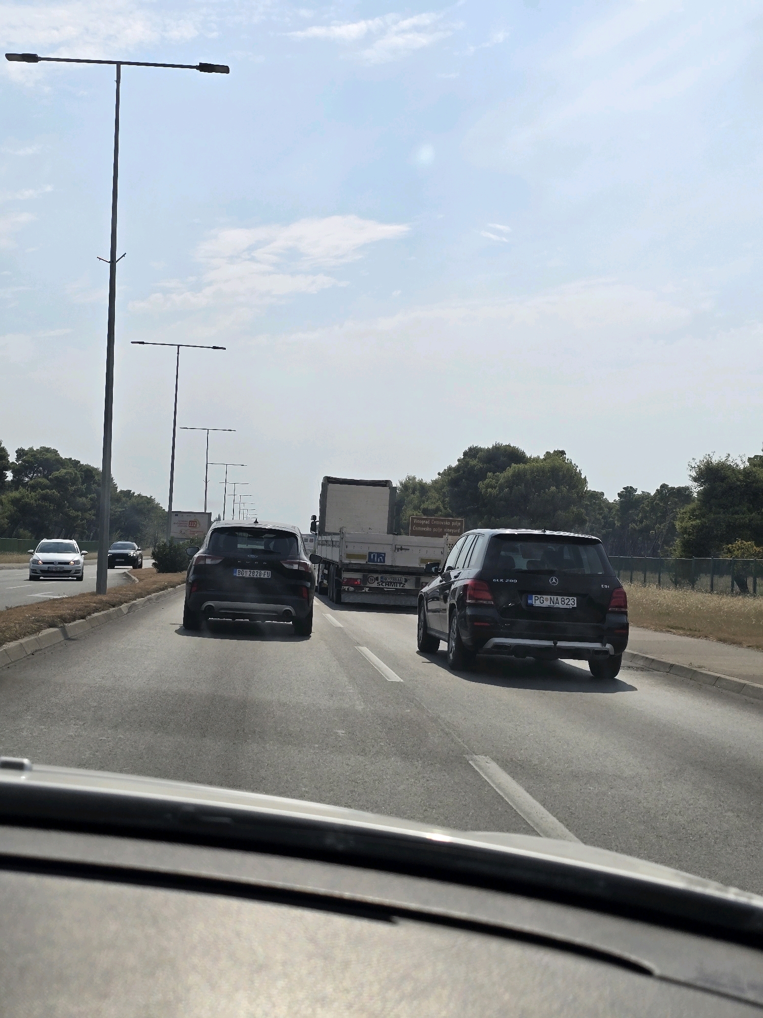 The image shows a view from inside a vehicle, looking forward onto a multi-lane road. In the foreground, the dashboard of the vehicle is visible. On the road, there are several vehicles. A black SUV, possibly a Ford Kuga, with the license plate BG 2828 FU, is in the left lane. Ahead of it, in the right lane, is a black Mercedes-Benz SUV, with the license plate PG NA 823. Further ahead in the right lane is a large truck. In the left lane, further ahead, is a white Volkswagen Golf. The road is bordered by a sidewalk and vegetation, with trees lining the background. There are several streetlights visible along the side of the road. The sky is blue with some scattered clouds. The location is identified as the city of Podgorica, Montenegro.