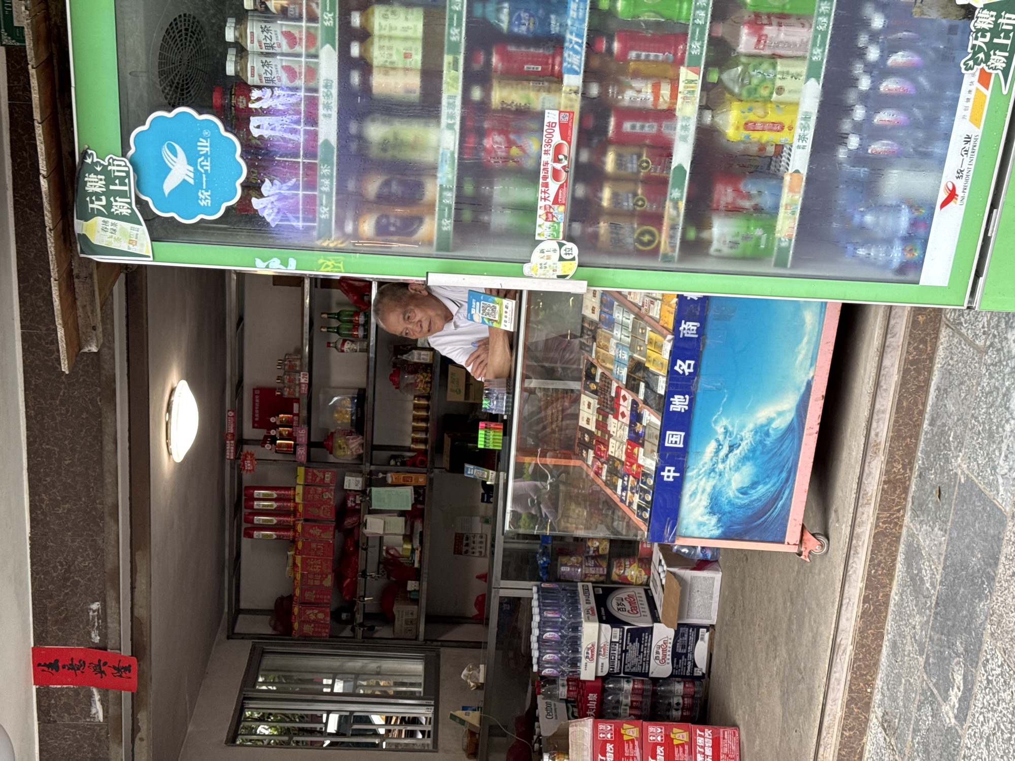 An older man is standing inside a small shop in Guilin, China. The shop is crammed with goods, including drinks in a refrigerator. The sign on the side says, "UNI-PRESIDENT ENTERPRISES." Other items include boxes of drinks and snacks, with a cooler stocked with various beverages visible through the glass door. The man is positioned behind a counter, presumably attending to customers. A bright light is visible on the ceiling inside the shop. The entrance area is a narrow space adjacent to a textured wall.
