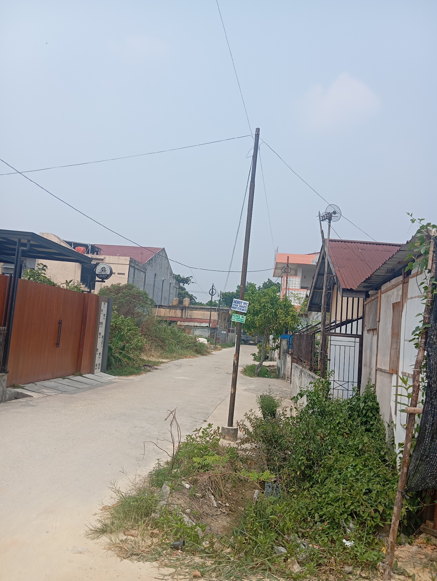 This is a ground-level, eye-level, medium shot image of a street scene in Pekanbaru, Indonesia. The image is taken on an overcast day, with a pale sky. The street is paved with concrete and is relatively narrow, with houses and buildings lining both sides.
On the left side of the street, there is a large, reddish-brown metal gate that appears to be the entrance to a property. To the left of the gate, there is a covered structure with a metal roof supported by poles, and part of a building with a tan facade. Behind the gate, dense green foliage is visible.
On the right side of the street, a residential area is depicted. There are several houses with various roof types, including corrugated metal and what appears to be tile. One building has a red corrugated metal roof and a white facade. Another building, further back, has an orange-tiled roof. A wooden utility pole with wires runs along the street, with some of these wires extending across the frame. There is also a smaller, dark-colored pole on the right side of the street.
Vegetation is abundant throughout the scene, with overgrown bushes and weeds growing along the sides of the street and in front of the houses. Some of the foliage is quite dense. In the middle ground, there is a tree with green leaves.
A sign on a utility pole on the left side of the street reads "SEDOT.WC HP. MANPEN 0821 7731 0031". Another sign, round and black with white text and a logo depicting a building, is visible on the left side of the street near the gate, it reads "AIRES VILLA".
In the distance, more buildings and trees can be seen. The overall impression is of a somewhat unkempt but lived-in urban or suburban environment.