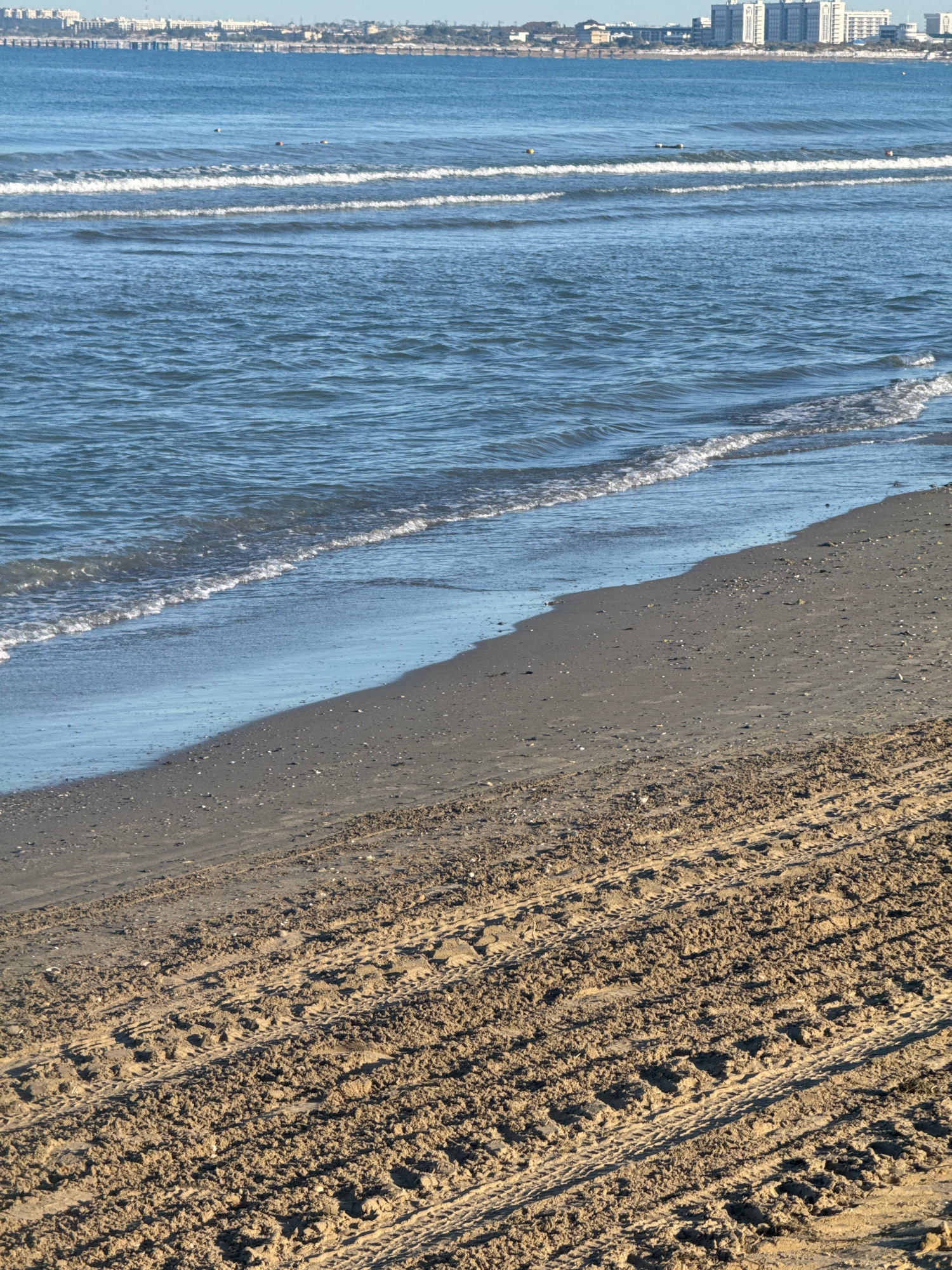 This is a photograph of a beach in Anapa, Russia, on a clear day. The foreground shows a sandy beach with tire tracks, indicating vehicle access. The middle ground features the gentle waves of the sea lapping onto the shore. In the background, across the water, is the Anapa skyline, characterized by several modern buildings and a distant city. There are no people visible in the frame. The scene depicts a tranquil coastal environment with evidence of human activity in the form of tire tracks on the sand.