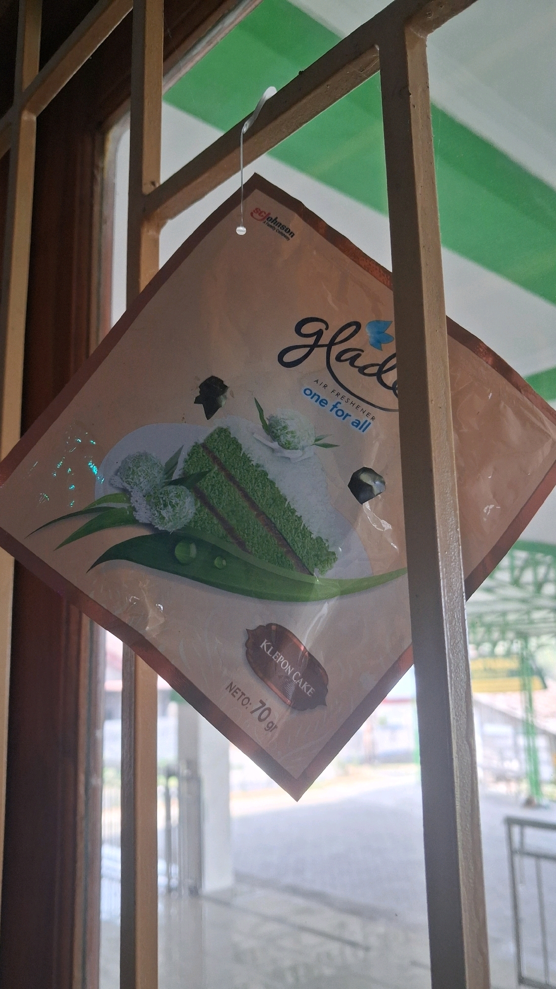 A Glade air freshener, depicting a slice of Klepon cake, is hanging on a wire hook from a metal grate. The grate appears to be part of a window frame, with a building and some greenery visible through it. The air freshener is diamond-shaped and positioned at an angle, showing the product's packaging with the brand name "Glade" prominently displayed. Below the cake image, "KLEPON CAKE" is written on a small label, followed by "NETO: 70 gr". The background suggests an outdoor environment, possibly in a residential or commercial area, given the presence of buildings and a paved surface. The overall lighting is somewhat diffused, indicating it might be daytime.