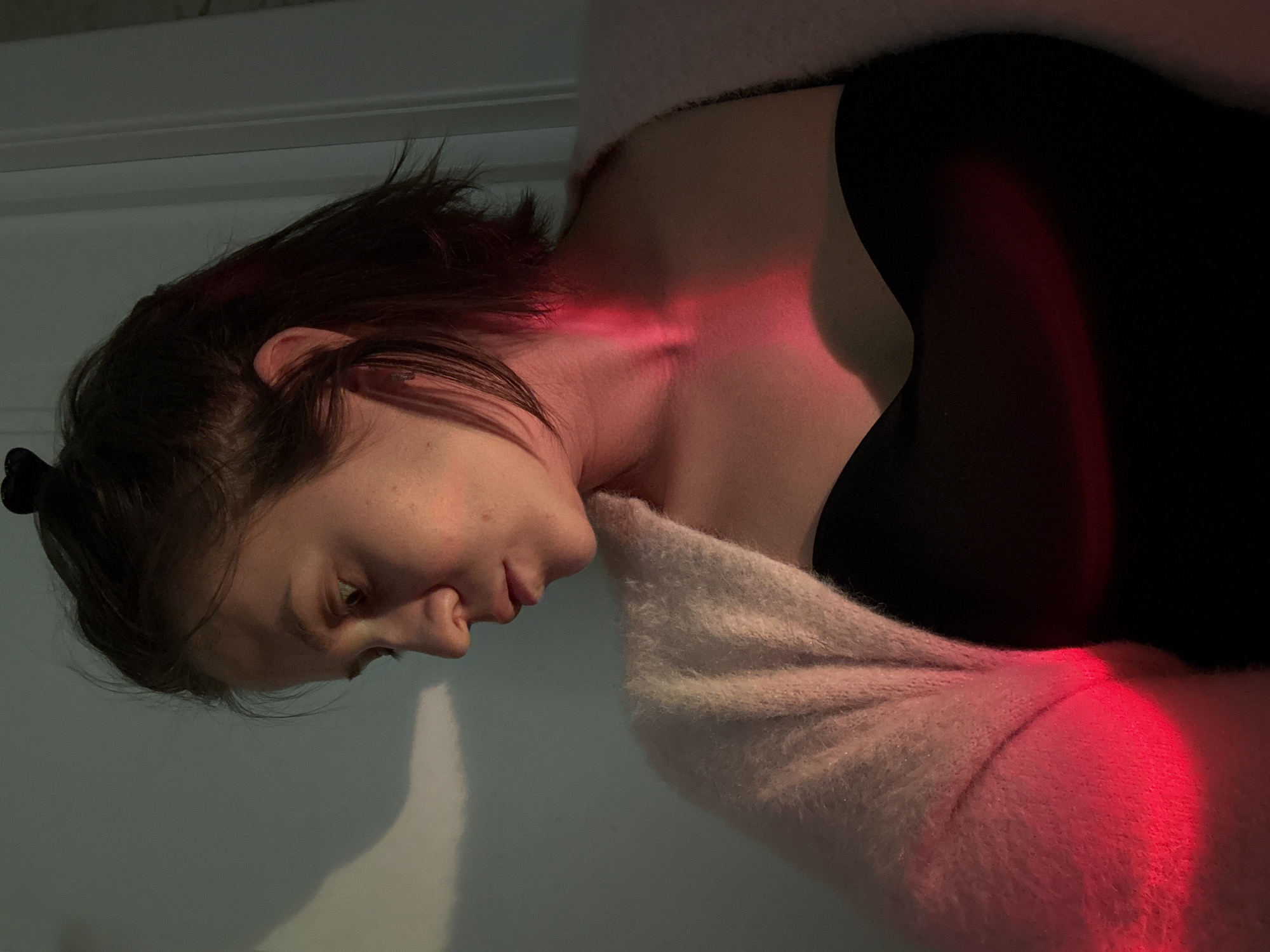 An indoor portrait taken in Pervomaysky, Russia, features a young adult individual, likely female, in a dim setting. The subject is seen in profile, looking downwards with a contemplative or melancholic expression. Her dark hair, which appears to have red or pink highlights, is pulled back from her face.
She is dressed in a dark top, possibly black, and a light-colored, fluffy sweater in a creamy or pale pink shade that covers her shoulders and arm. A striking red light illuminates her neck, collarbone area, and parts of her arm and sweater, casting dramatic shadows and highlights across her features and clothing. This artificial lighting creates a moody and intimate atmosphere.
The background is a plain, light-colored wall, with a subtle horizontal line, possibly a door frame or molding, visible in the upper part of the image. The overall ambient light is low, emphasizing the effect of the localized red illumination. No specific activity other than posing is depicted. There is no visible text within the image.