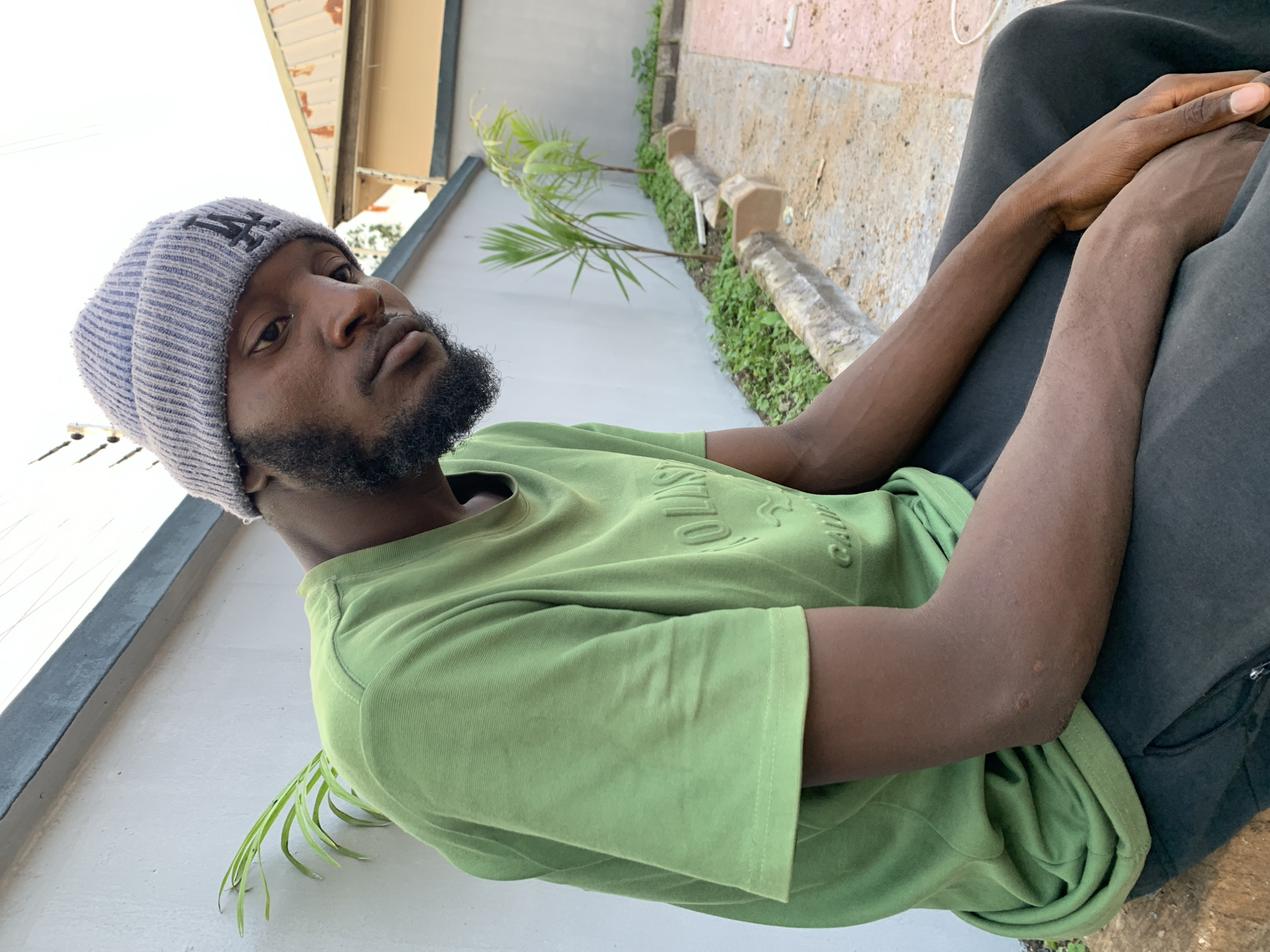 In Bukuru, Nigeria, a young Black man with a beard and a grey beanie sits outdoors, leaning against a light-colored wall. He is wearing a bright green t-shirt with embossed lettering and dark pants. Two small palm trees are planted in a garden bed behind him. The ground in front of the garden is paved with concrete and some sand. A building with a tan roof is visible in the background.