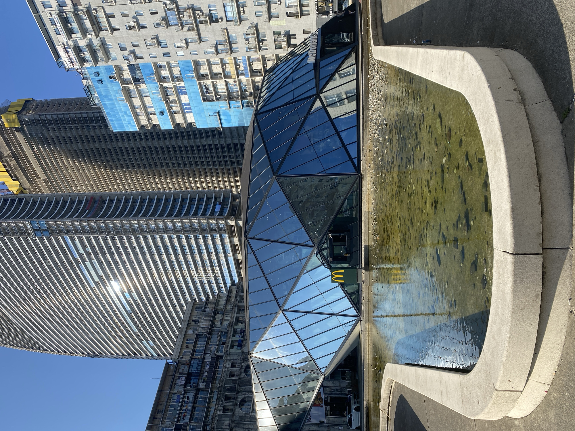 The image captures a modern architectural scene in Batumi, Georgia, featuring a geometric glass building with a McDonald's restaurant. The building's facade is composed of numerous triangular glass panels, reflecting the bright blue sky and surrounding structures. In front of the glass building, a shallow, rectangular fountain with a concrete border contains clear water with scattered pebbles and some debris. A prominent golden McDonald's logo is visible on a freestanding sign. Behind the glass structure, a tall, unfinished skyscraper with exposed concrete framework stands under construction, alongside other apartment buildings of varying architectural styles, including one with a distinctive blue glass facade. The overall environment suggests an urban setting with a mix of new and ongoing construction.