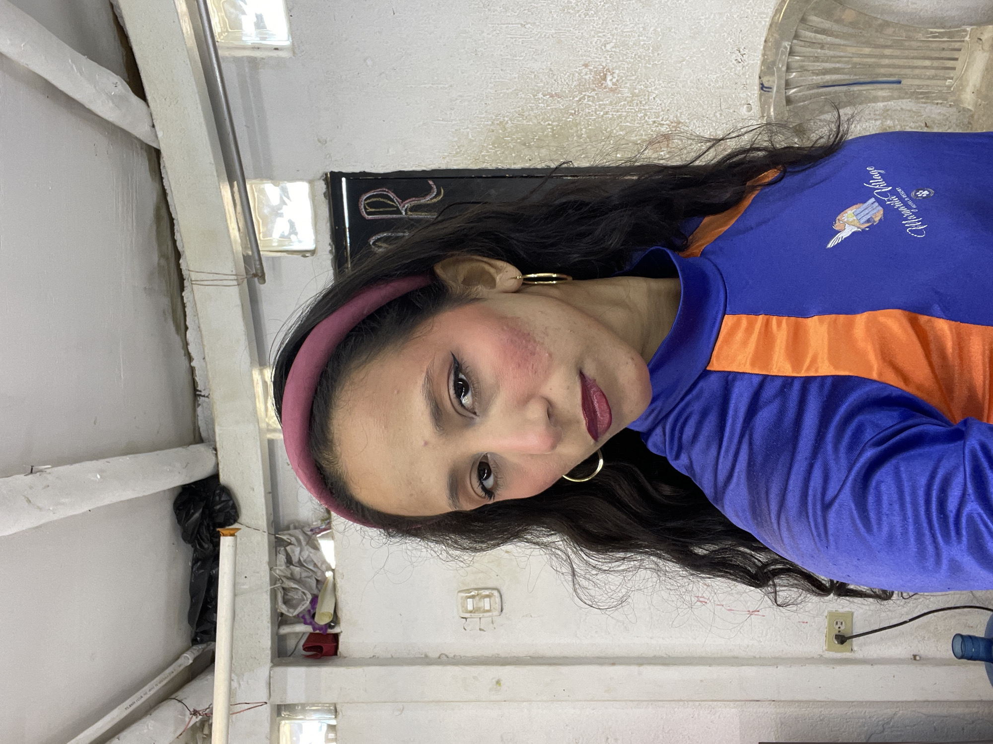 A woman with long, dark, wavy hair and a pink headband stands in front of a white, textured wall in what appears to be an indoor, possibly unfinished or utility space. She is wearing a blue shirt with orange accents and a logo on the left side that reads "Margarita Village Hotel & Resort". Her makeup includes dark eyeliner, eyeshadow, and a dark lipstick. She is wearing large hoop earrings. The background shows exposed pipes and electrical conduit on the ceiling, as well as some storage items on a shelf, including a black garbage bag. A partially visible chair is on the right side of the image. The overall environment suggests an informal or backstage setting, possibly in Porlamar, Venezuela, given the hotel logo.