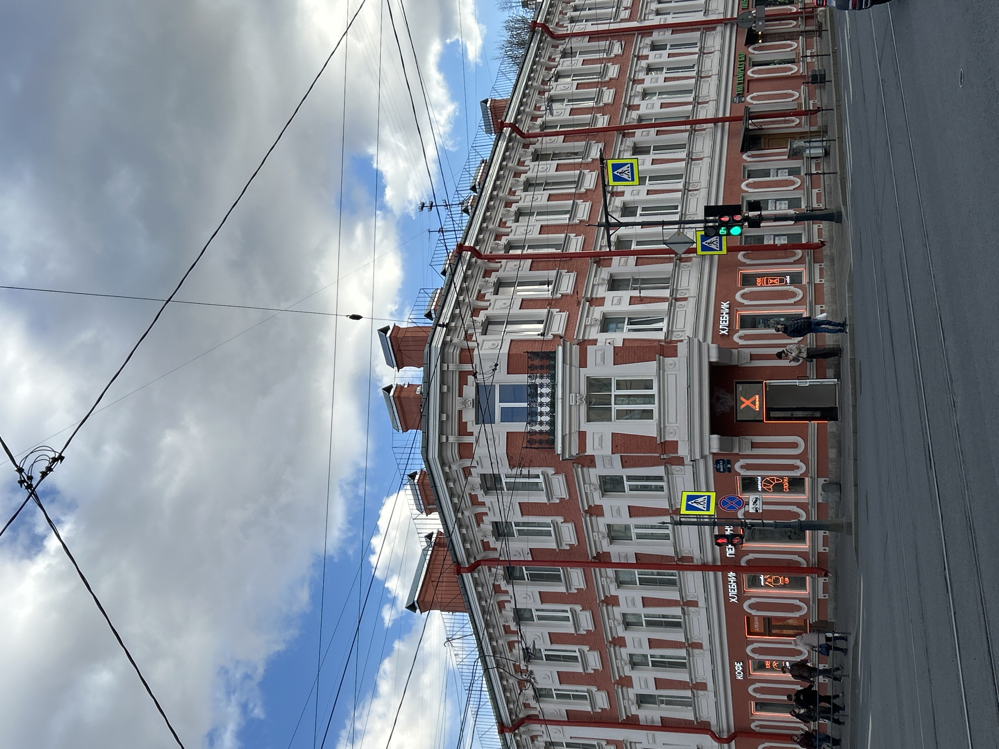 The image, when properly oriented, presents a vibrant daytime street scene in Saint Petersburg, Russia. A grand, multi-story historical building with a distinct red brick facade and intricate white decorative elements dominates the right side. The building features numerous windows, some adorned with small balconies and wrought-iron railings. Red drainpipes extend vertically down the facade. The rooftop is partially visible, showing several small structures and a metal railing.
At ground level, the building houses various businesses, indicated by brightly lit signs. A wide asphalt street with visible tram tracks runs parallel to the building. Several pedestrians can be seen on the sidewalk, casually strolling, with a couple more prominent in the foreground.
The urban environment includes essential street furniture: traffic lights displaying both green and red, yellow diamond-shaped pedestrian crossing signs, and other road signs. A network of overhead wires, likely for trams or trolleybuses, crisscrosses the sky. The sky itself is a mix of light grey clouds and clear blue patches, suggesting a partly cloudy day.
Visible text on the storefront signs is in Russian. "ХЛЕБНИК" (Khlebnik), likely a bakery or cafe, appears multiple times. Other signs read "КОФЕ" (Kofe - Coffee), "ПЕКАРНЯ" (Pekarnya - Bakery), "СДОБА" (Sdobа - Sweet pastry/bun), and "ТВОРОГ" (Tvorog - Cottage cheese). One prominent sign displays a large "X" logo. Another sign indicates "ПИВНОЙ БАР" (Pivnoy Bar - Beer Bar) and "ПОДВАЛ" (Podval - Basement). The architecture and Russian text firmly place the scene in Saint Petersburg.