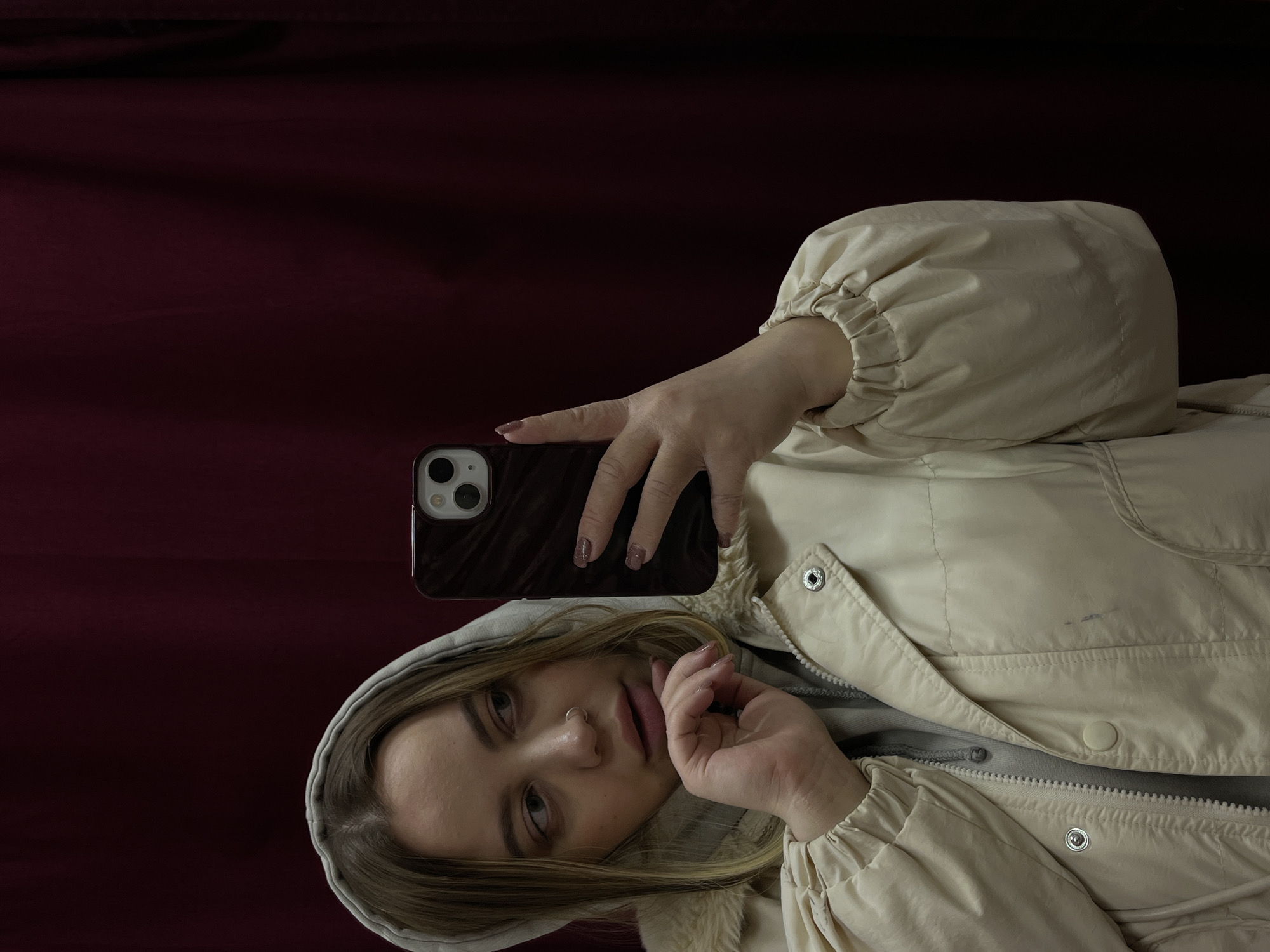 A woman in a light-colored hooded jacket is taking a selfie with her phone. The phone has a dark brown case. The background is a dark red fabric. The image does not contain information about location in the city of Novopokrovskaya, Russia.