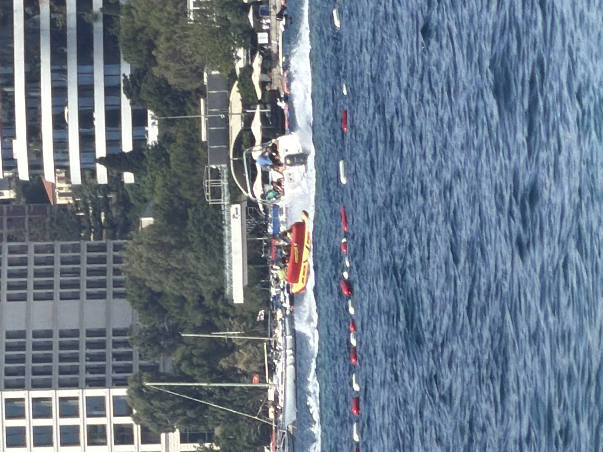The image captures a coastal scene in Boreti, Montenegro, with a high-angle perspective. The dominant feature is the blue sea occupying the right half, marked by a line of red and white buoys. Adjacent to the shoreline is a beach area with sun umbrellas and a boat towing a yellow inflatable. People are visible on the boat and beach. To the left of the beach, there is dense greenery and tall buildings, one characterized by repeating window units. The overall scene suggests a leisure or tourist activity.