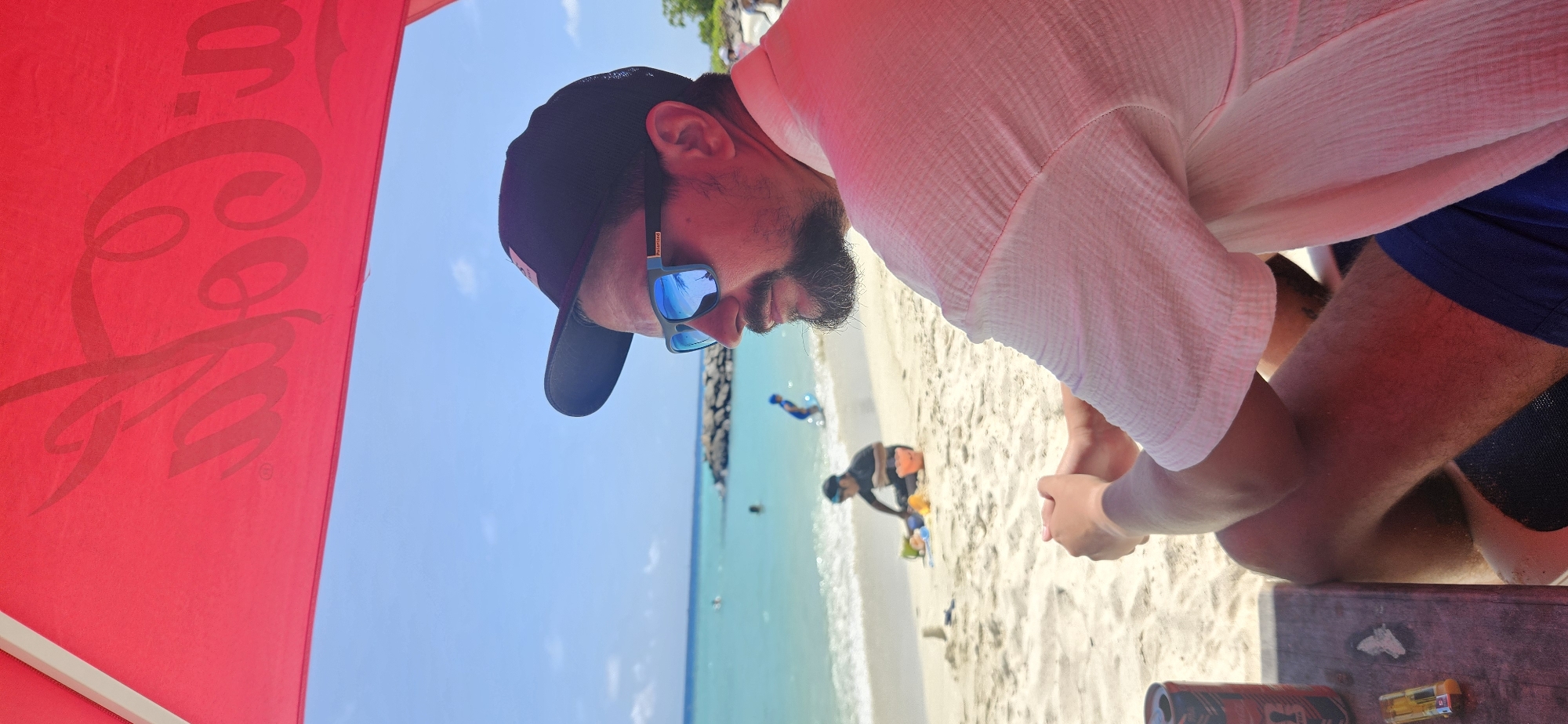 A man wearing a black baseball cap and blue-tinted sunglasses sits under a red umbrella, looking out at a tropical beach. The umbrella has a Coca-Cola logo visible on its underside. The man has a beard and is wearing a light-colored, crinkled short-sleeved shirt and blue shorts. His hands are clasped in his lap. To his left, on a wooden surface, are a red and silver can and a yellow lighter. The background shows a sandy beach with people in the distance. Two individuals are visible near the water, one appearing to be a child in blue swimwear standing in the shallow waves, and another person in dark clothing sitting on the sand playing with colorful toys, possibly a bucket and spade. The ocean is a clear turquoise, and beyond it, a rocky breakwater is visible. The sky is bright blue with a few wispy clouds. The scene is consistent with a beach environment on Maafushi Island, Maldives, known for its tourist resorts and clear waters.
