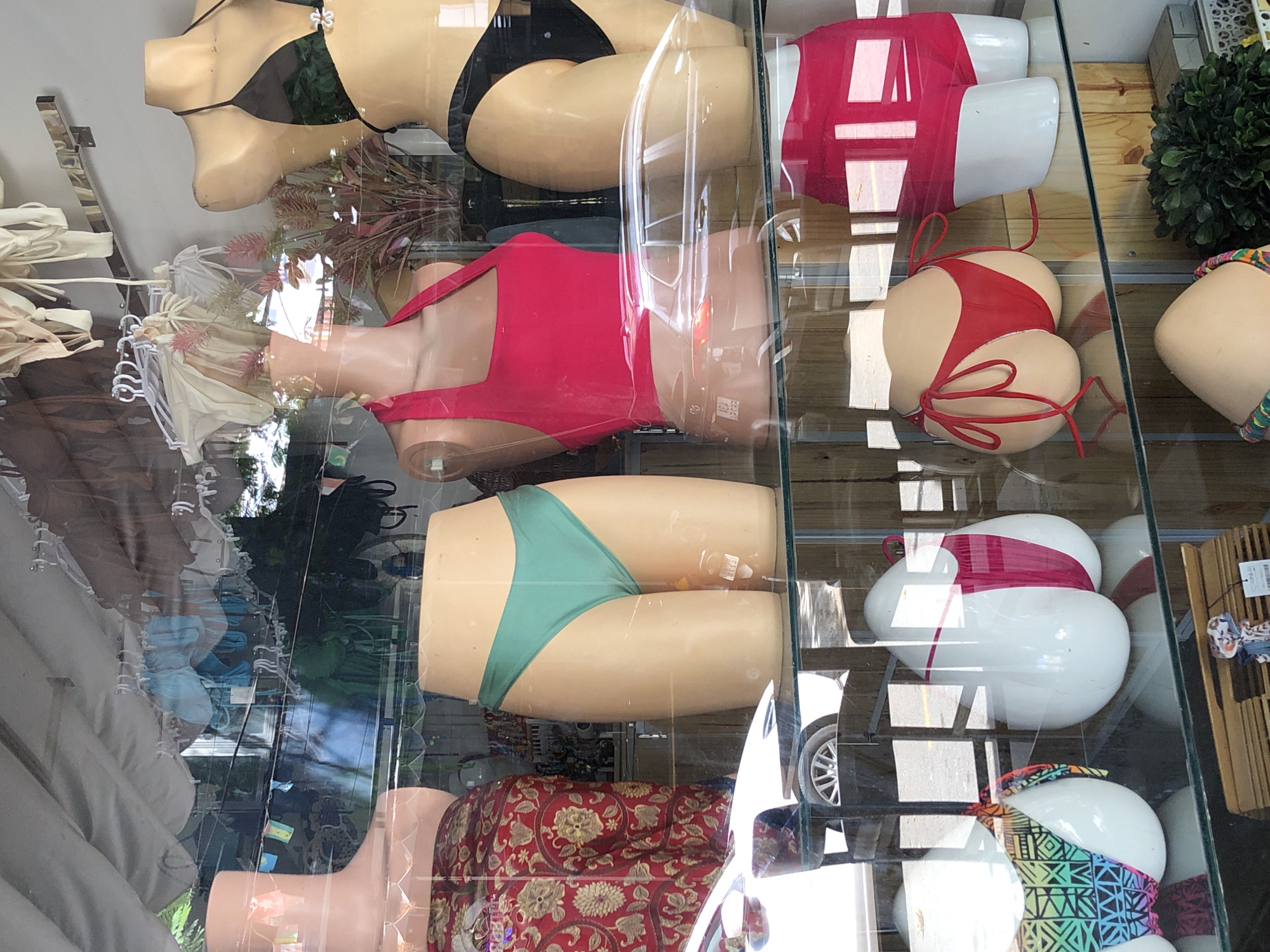 The image shows a display window of a store. There are mannequins dressed in swimwear. In the foreground, from left to right, are a mannequin torso with a patterned skirt, a mannequin lower body in a green bikini bottom, a mannequin torso in a pink top, and a mannequin lower body in a black bikini bottom. Below these, there are two white, egg-shaped mannequins wearing bikini bottoms, one with a multi-colored geometric pattern and the other with a pink and white design. To the right of these, there is a mannequin lower body in a red and white bikini. In the background, there are racks of swimwear, some plants, and reflections of the street outside, including a white car. The store appears to be a boutique selling swimwear. The scene is consistent with a retail establishment in Đà Nẵng, Vietnam, given the likely climate and fashion trends.