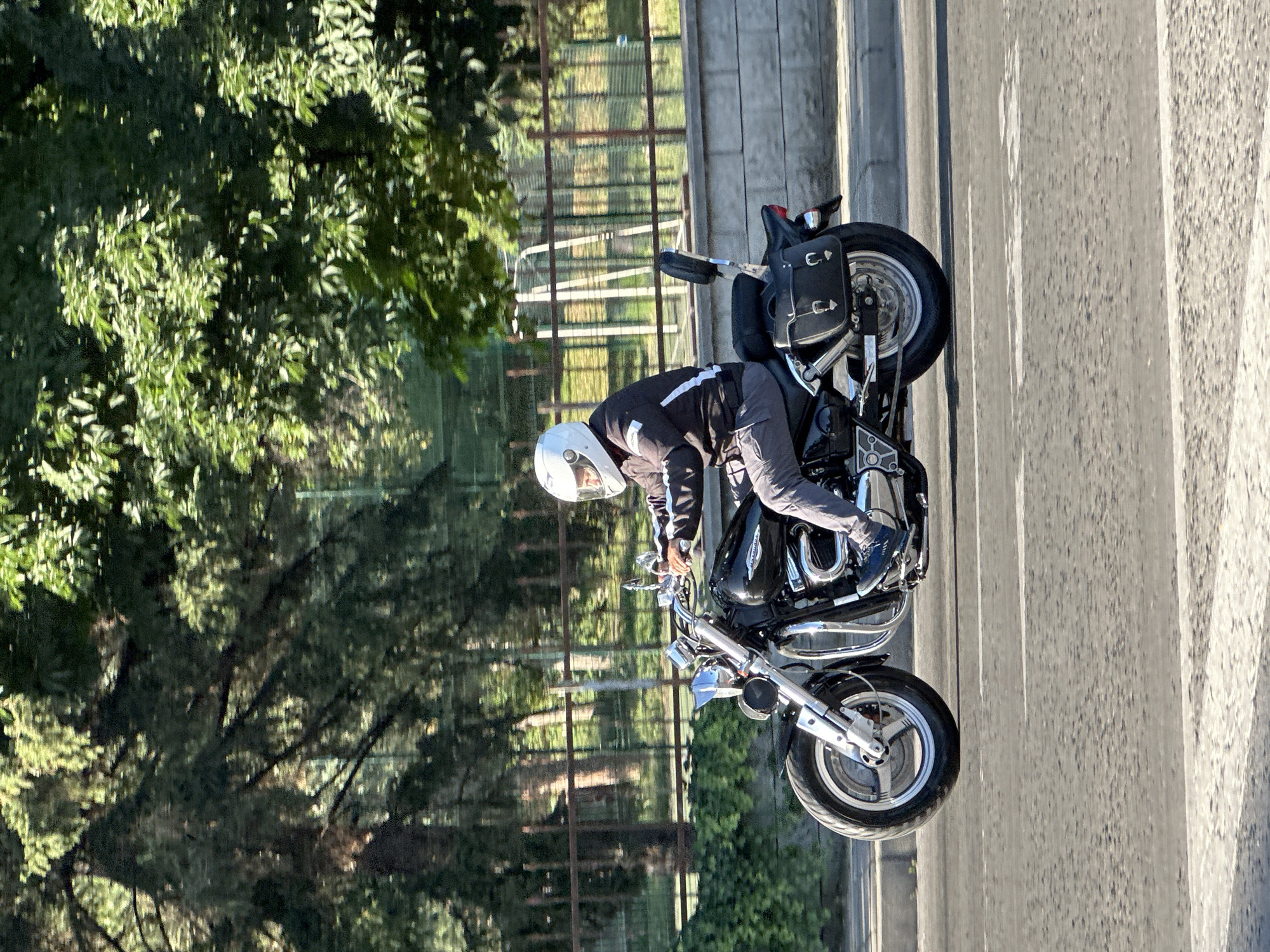The image features a person on a motorcycle, positioned on a street. The person is wearing a white helmet and riding gear, with a black and white jacket and gray pants. They appear to be leaning forward, possibly adjusting something on the motorcycle. The motorcycle is black with chrome accents and saddlebags. In the background, there are trees with green foliage, a fence, and a wall or building with vertical lines. The scene suggests an outdoor setting, possibly during the daytime given the lighting. The image is oriented vertically, with the motorcycle and rider appearing on the right side of the frame, and the greenery on the left. The ground is paved, indicating an urban or suburban street. The overall impression is a candid moment of a motorcyclist on the road.