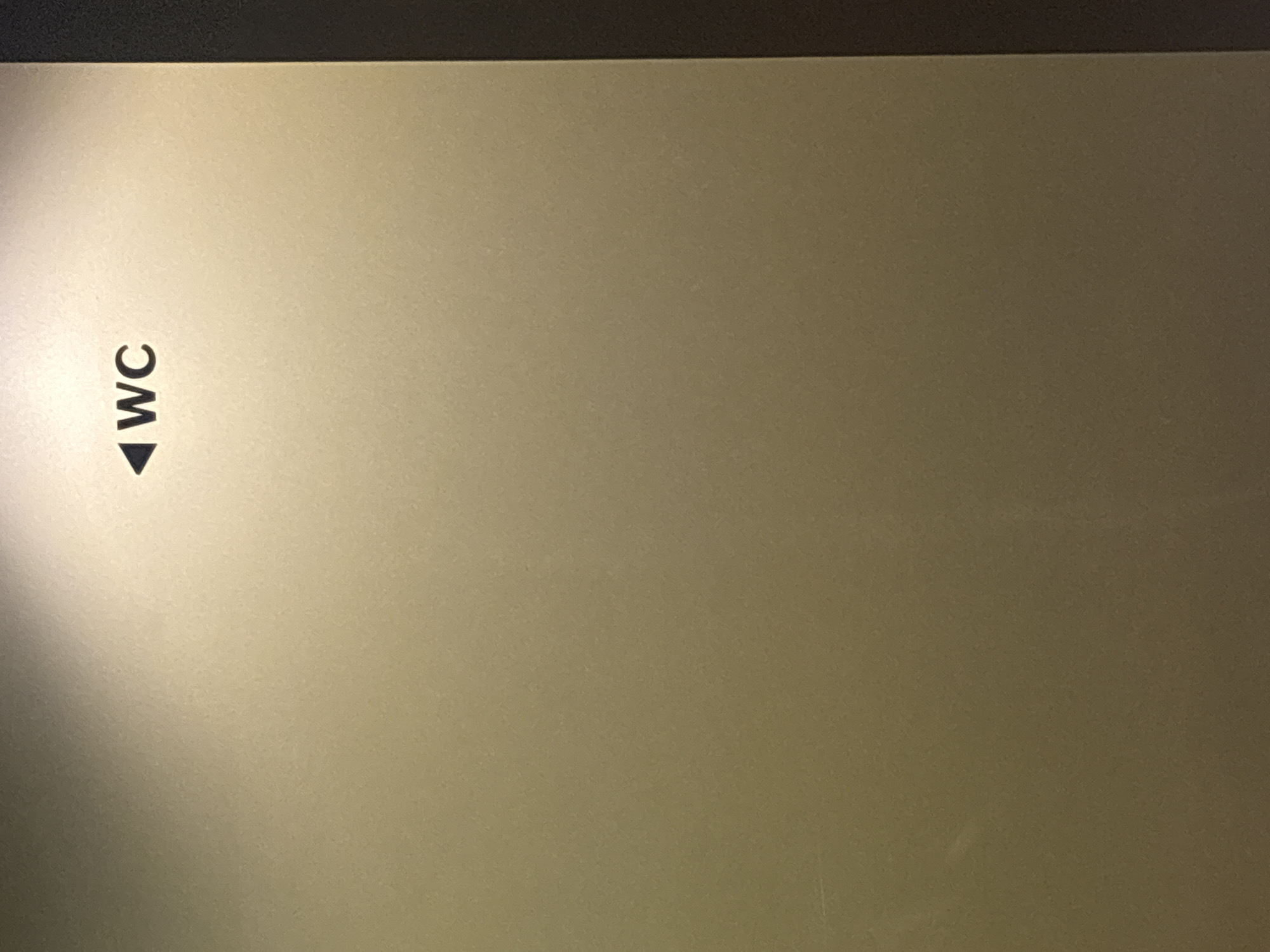 The image shows a sign with the text "WC" and an arrow pointing left. The sign is black and is placed on a beige metallic surface. The lighting suggests it is indoors, likely in a public space such as a restaurant, hotel, or airport. The specific location in Burgas, Bulgaria is not discernible from the image.