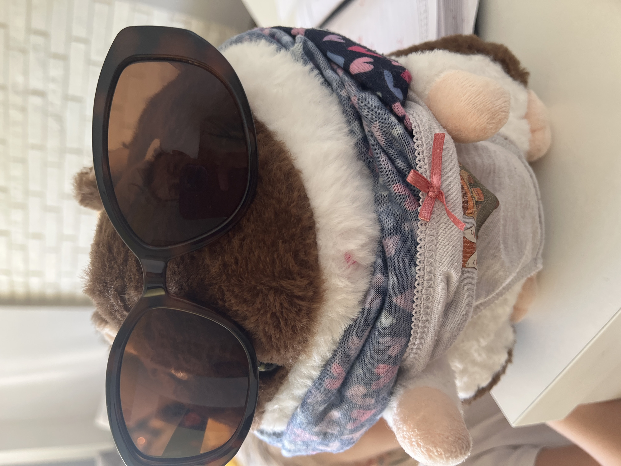 A stuffed animal, likely a guinea pig, wears brown sunglasses and several layers of clothing, including a gray t-shirt and a blue floral scarf. The animal rests on a white surface. The location is unknown.