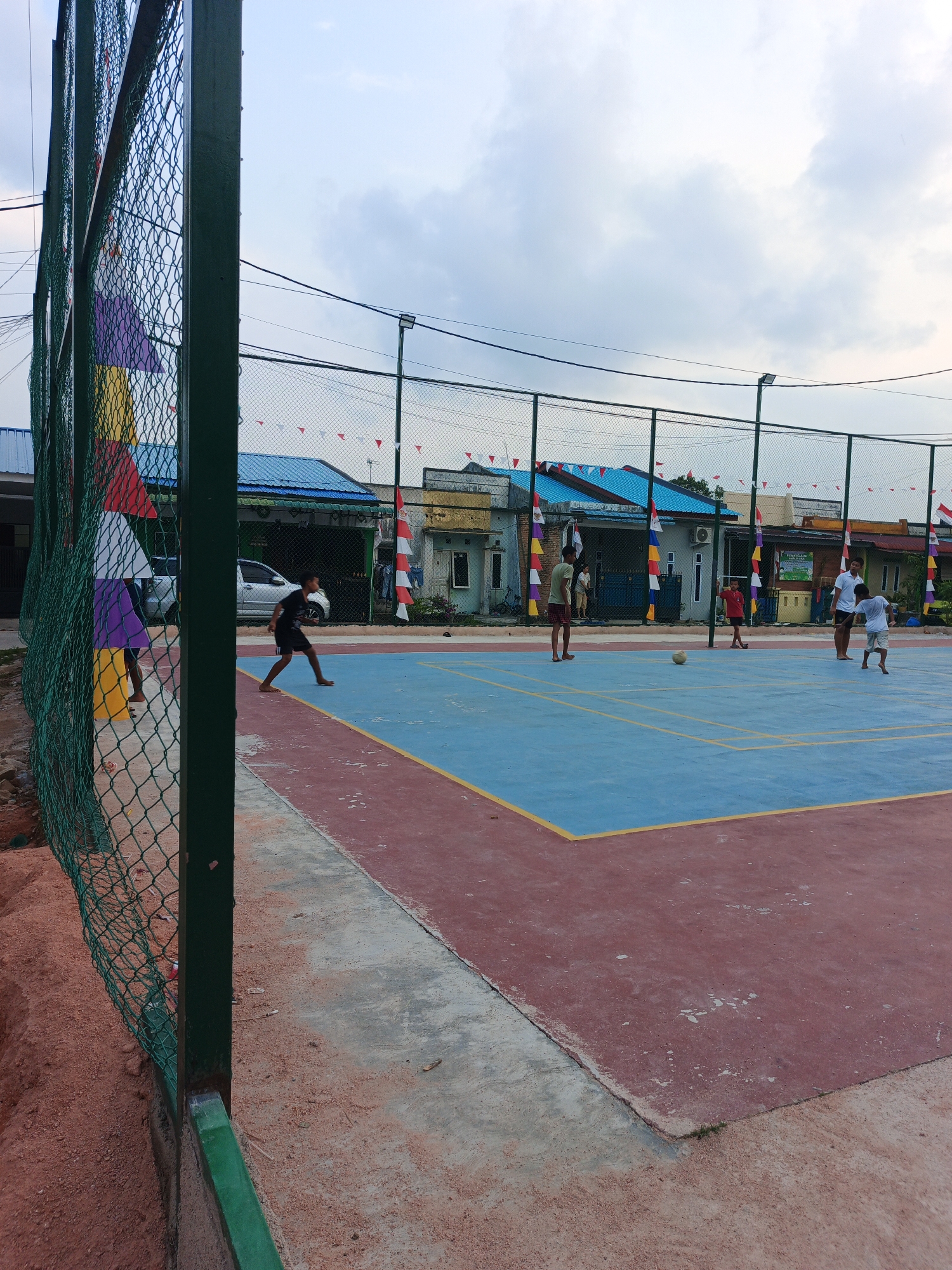 This is a medium-angle, eye-level, outdoor, full-shot photograph of a volleyball court in Batam, Indonesia. In the foreground, a green metal fence with wire mesh encloses a sandy area. Beyond the fence, a blue and red volleyball court with yellow markings is visible. Several individuals are playing volleyball on the court. In the background, residential buildings with blue roofs, decorated with red and white flags and colorful banners, are present. The sky is cloudy and overcast.