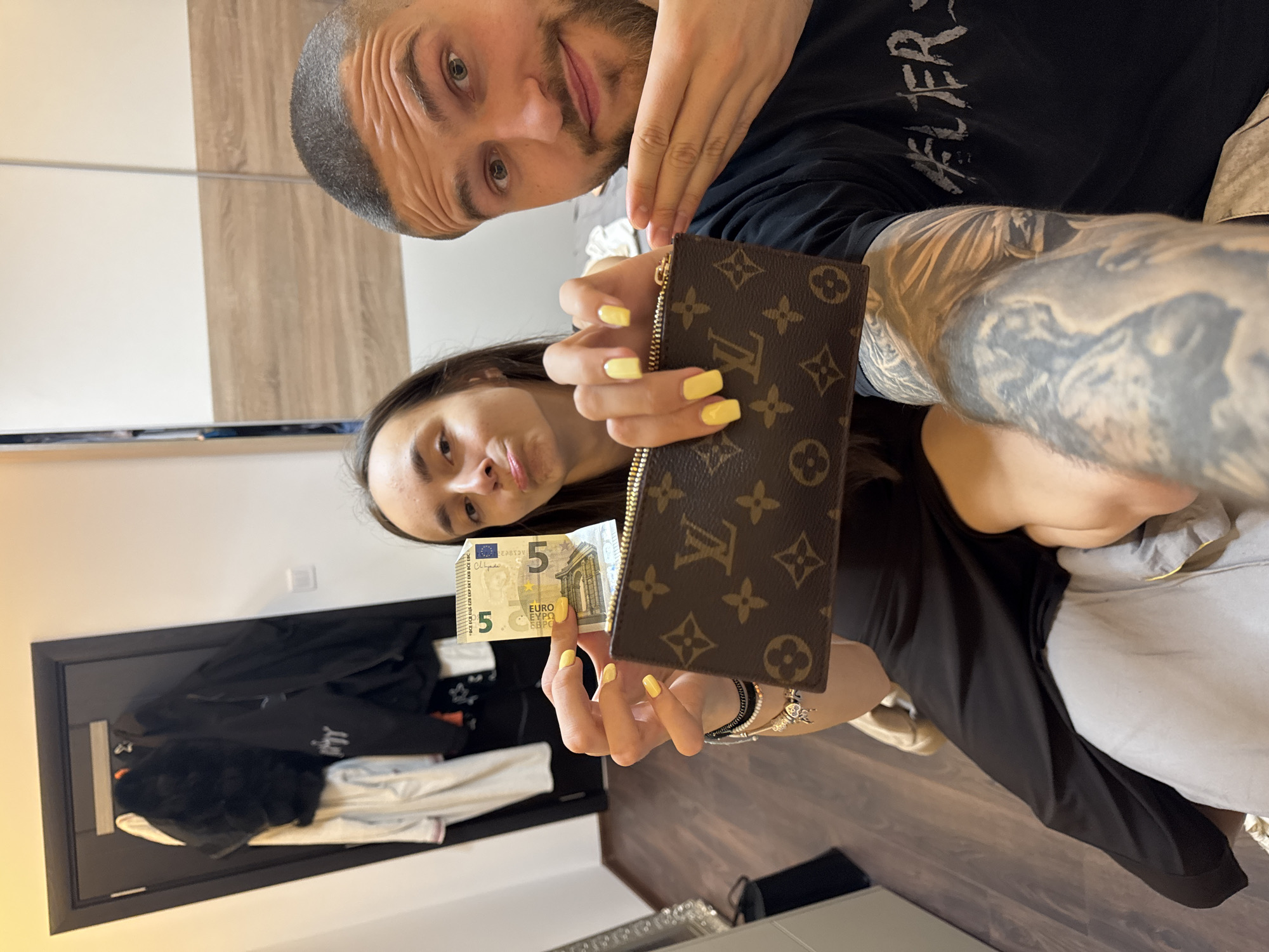 A selfie features a man and a woman inside what appears to be an apartment or house. The image is oriented sideways. The man, with short hair and a beard, is in the upper portion of the frame. He is partially visible, with only his face, part of his arm, and chest shown. He is wearing a black t-shirt with some white lettering.
The woman, who appears to be holding the camera for the selfie, is positioned below him. She has long brown hair and is holding a brown, Louis Vuitton-branded coin purse in her right hand and a 5 Euro bill in her left hand. Her fingernails are painted a bright yellow. She is wearing a black dress and a wrist full of bracelets.
The background includes a closet door with jackets and clothes hanging inside. The walls are painted white and beige. The flooring appears to be hardwood.
The location and city, Sofia, Bulgaria, cannot be determined from the image.