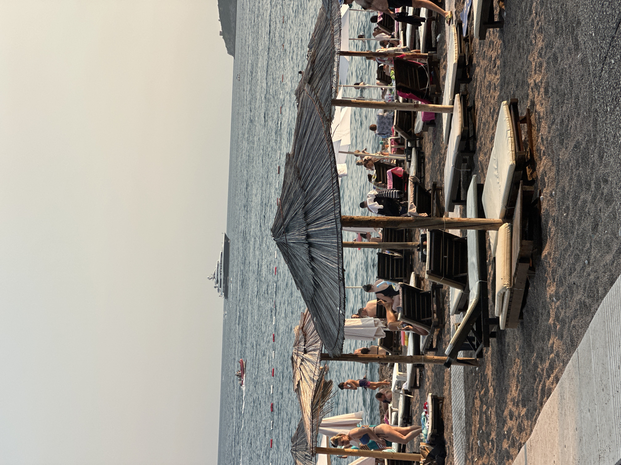 The image depicts a sun-drenched beach scene in Boreti, Montenegro. The foreground is dominated by rows of empty lounge chairs and thatched umbrellas set against a pebbly sand beach. A concrete walkway runs along the left side of the frame. In the middle ground, beachgoers are visible, some reclining on chairs, others wading in the calm, blue water. A few individuals are standing and interacting near the water's edge. The background features the vast expanse of the Adriatic Sea, with a large yacht sailing in the distance and a smaller red boat closer to shore. A hilly coastline with a prominent structure on it is visible on the right horizon. The sky is a pale, hazy blue, indicating a warm, possibly late afternoon, setting.