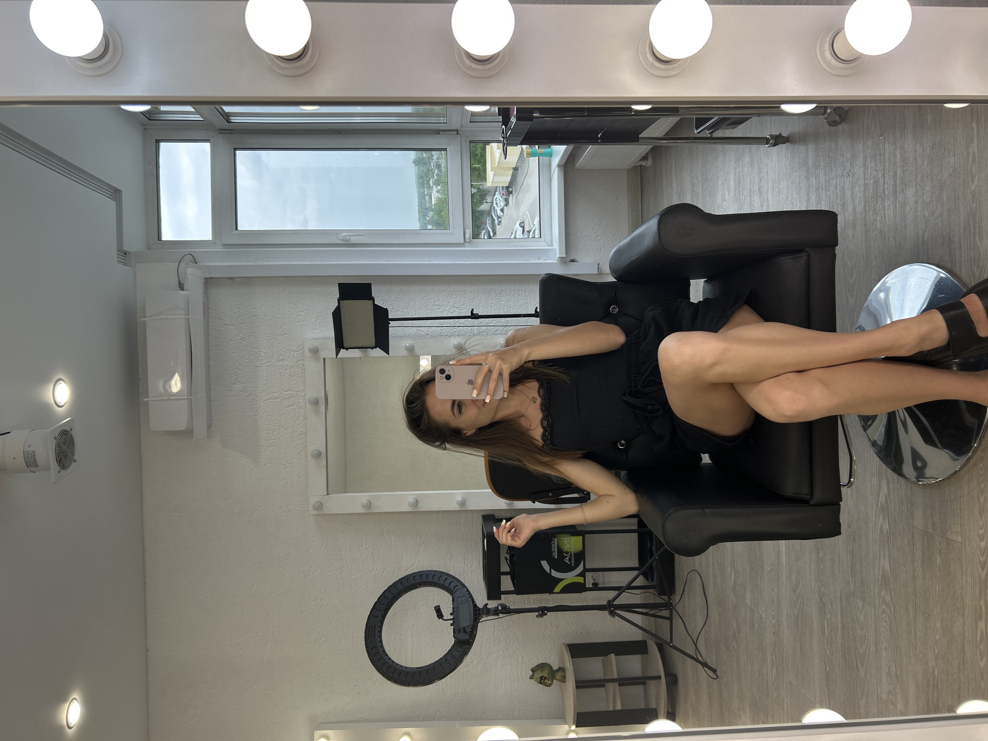A young woman with long brown hair, wearing a black lace-trimmed slip dress, is seated in a black leather chair in what appears to be a salon or dressing room. She is holding a pink smartphone, taking a selfie in a large mirror that is part of a vanity with illuminated bulbs. The mirror also reflects a window showing an overcast sky and a view of a street with parked cars and buildings. To the left of the woman, a ring light on a stand is visible, along with a small side table holding a decorative item. The room has white textured walls and a light-colored wood floor. In the background, equipment like a portable air conditioner unit and a makeup artist's cart are present.