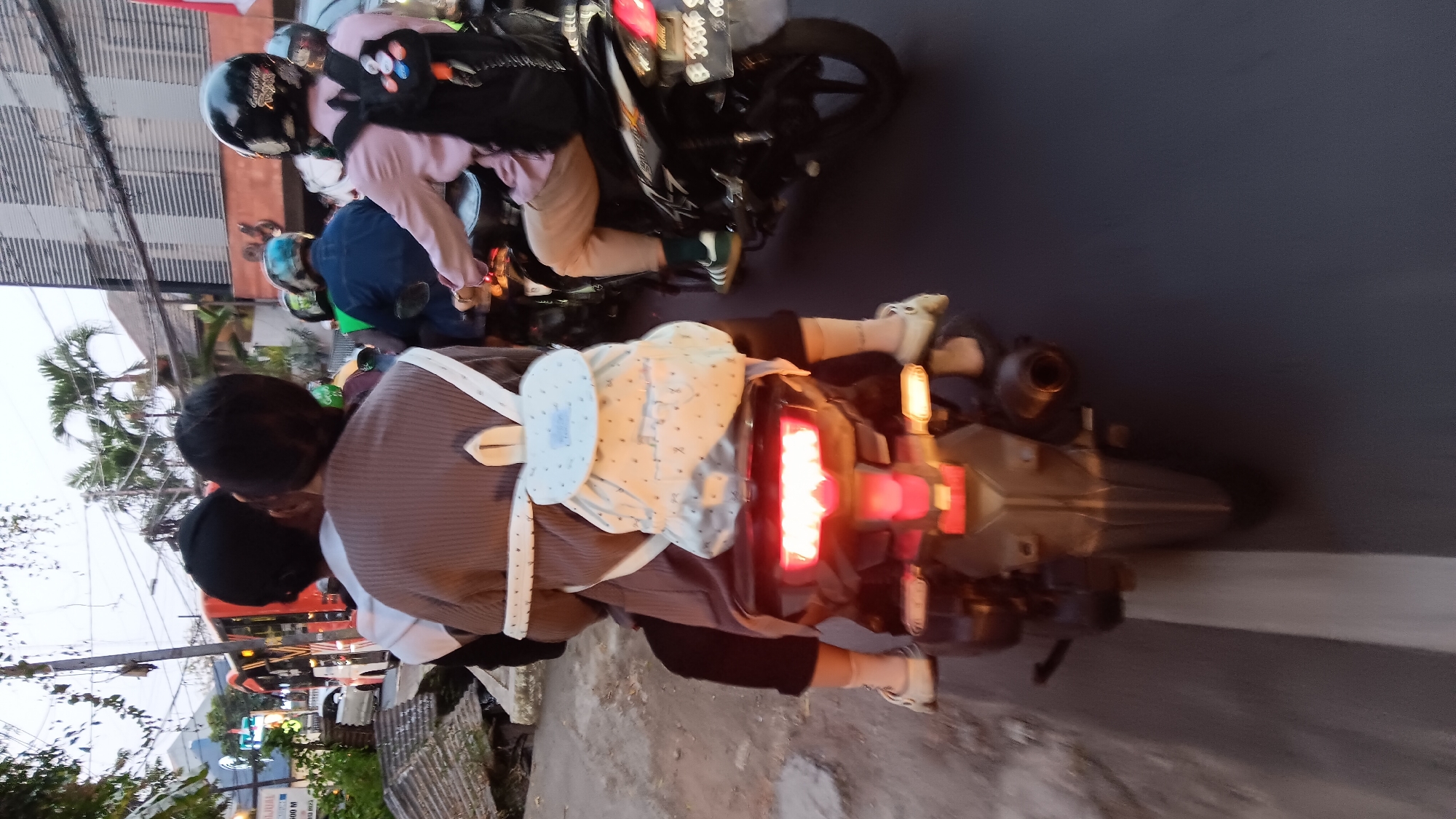 The image shows a group of people riding motorbikes on a street in South Jakarta, Indonesia. In the foreground, two individuals, possibly a parent and child, are riding a motorbike with the person in the back wearing a brown sweater and a light-colored backpack with a star pattern. The motorbike's taillights are illuminated. To the right, another person is riding a motorbike, wearing a pink shirt, beige pants, and a black backpack. Several other motorbikes with riders wearing helmets are visible in the background, suggesting a traffic scene. The environment includes buildings, palm trees, and overhead wires, typical of an urban setting. The lighting suggests it is either early morning or late afternoon.