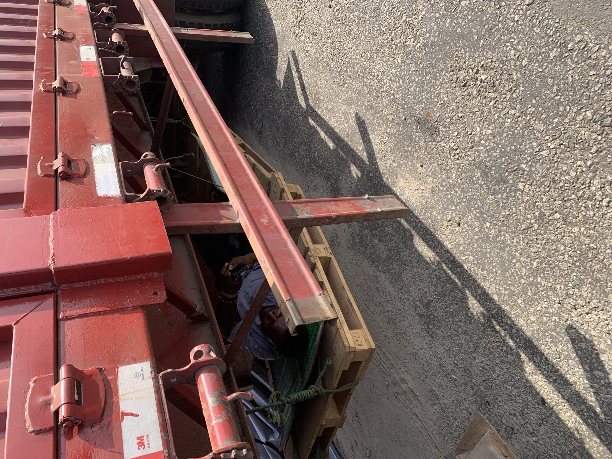 A top-down, angled view shows a red shipping container and a pallet, with a dark-skinned man partially visible underneath. The man appears to be lying down. The container is secured with a metal frame and various mechanical components. The setting appears to be an urban or industrial area with an asphalt surface. The location is Adankolo, Nigeria.