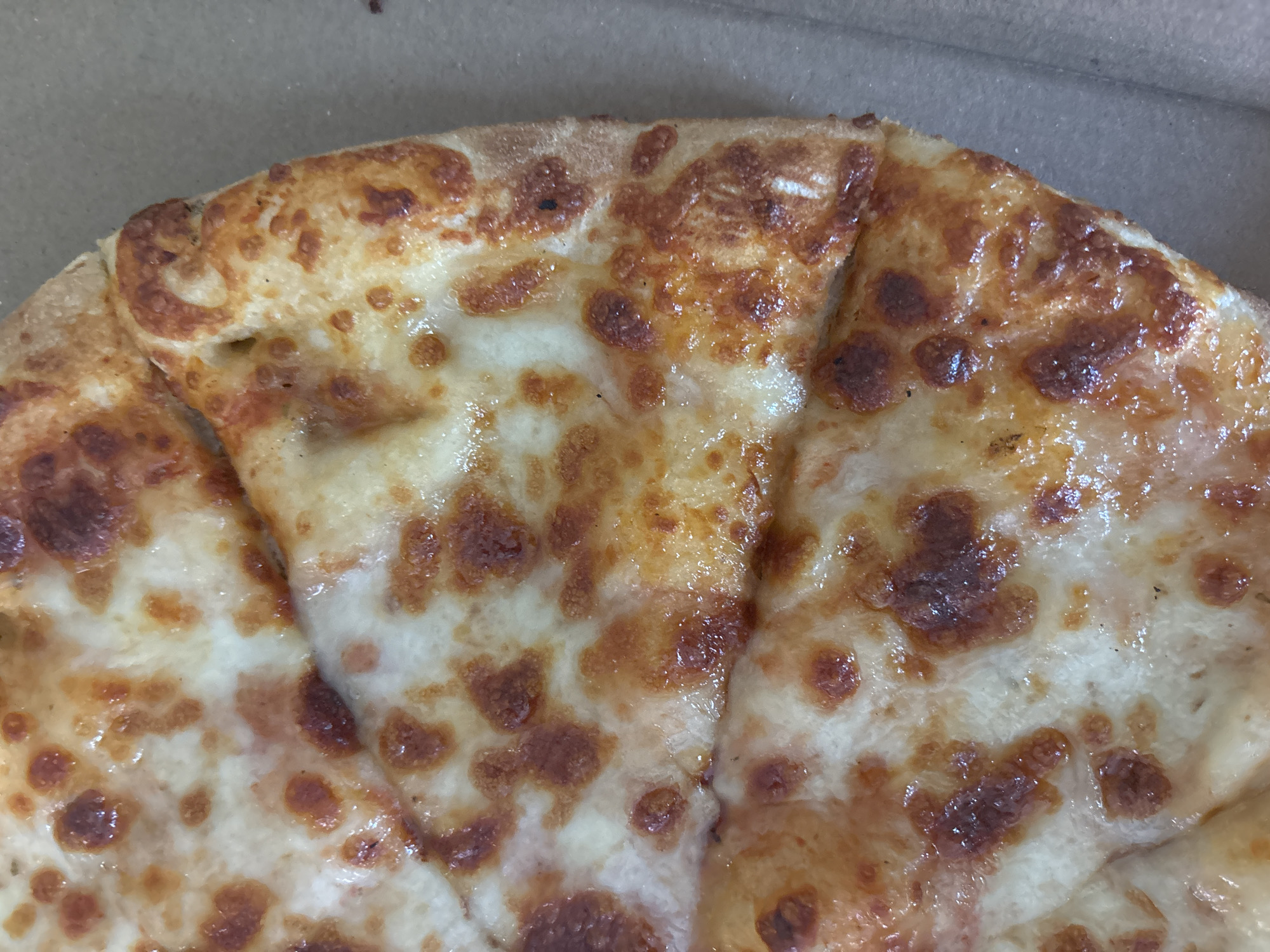This media file presents a close-up, overhead view of several slices of cheese pizza nestled within a brown cardboard pizza box. The primary subject is the pizza, which is generously covered in melted, bubbly cheese. The cheese displays various shades of golden-brown caramelization and some darker, charred spots, indicating it has been recently cooked. A subtle sheen across the cheese suggests the presence of oil or moisture. The visible crust edges are golden brown. The background and parts of the foreground reveal the plain, light brown interior of a standard pizza box. There are no people visible, nor is any specific activity taking place other than the static presentation of the pizza. No text is visible within the image. While the image itself provides no direct cues for time of day or weather, the context places the scene in Sofia, Bulgaria.