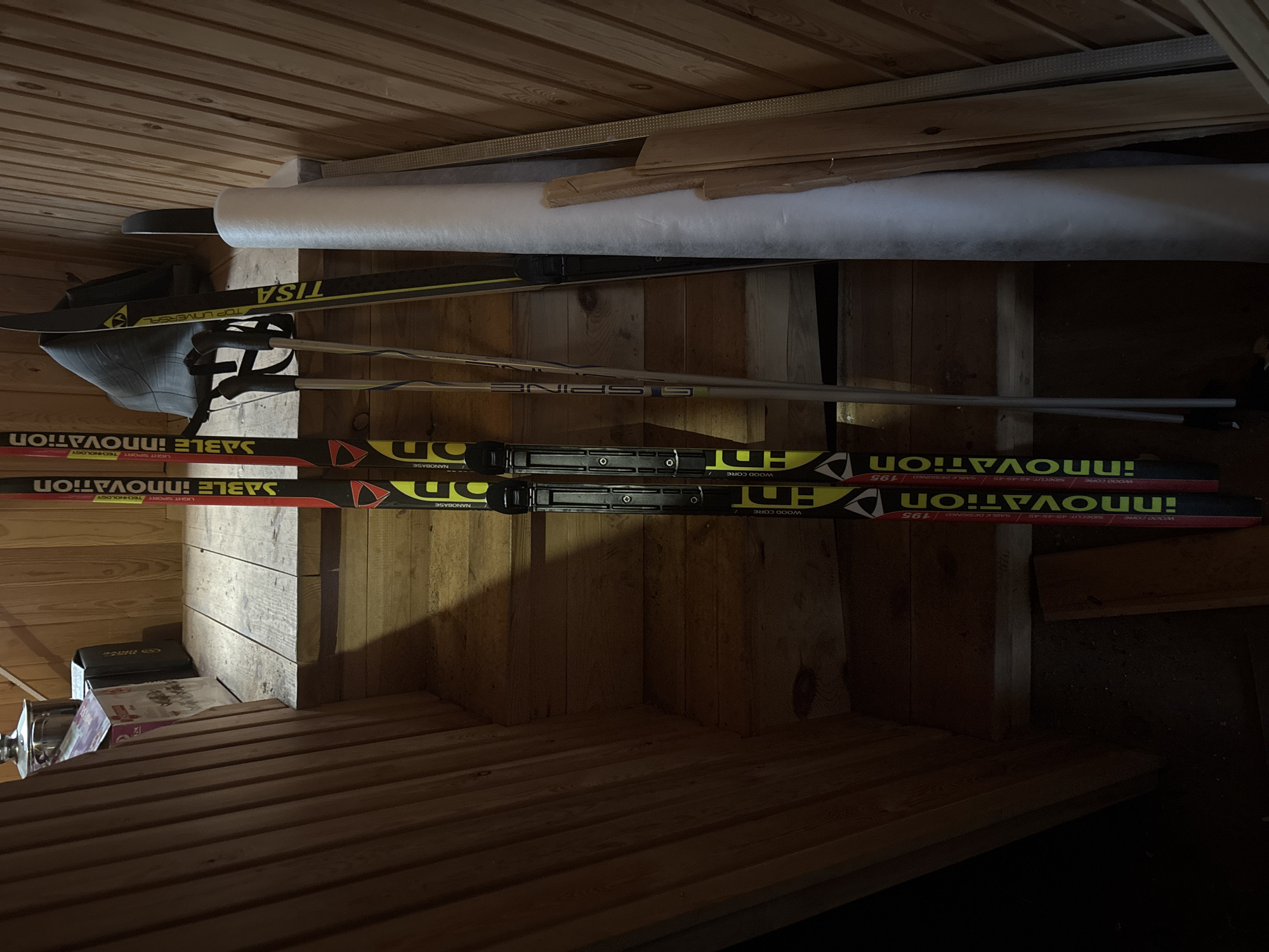 The image shows a storage area, possibly in a house or cabin, with a wooden ceiling and walls. A pair of cross-country skis with the brand name "Sable Innovation" are propped against the wall, along with ski poles and another pair of skis labeled "Tisa Top Universal." A roll of white material is placed above the skis. Below, various boxes and containers sit on a wooden shelf or platform. The image appears to have been taken from a low angle, looking upwards into the storage space. There is no indication of the city of Zagatuy, Russia, or any people in the image.
