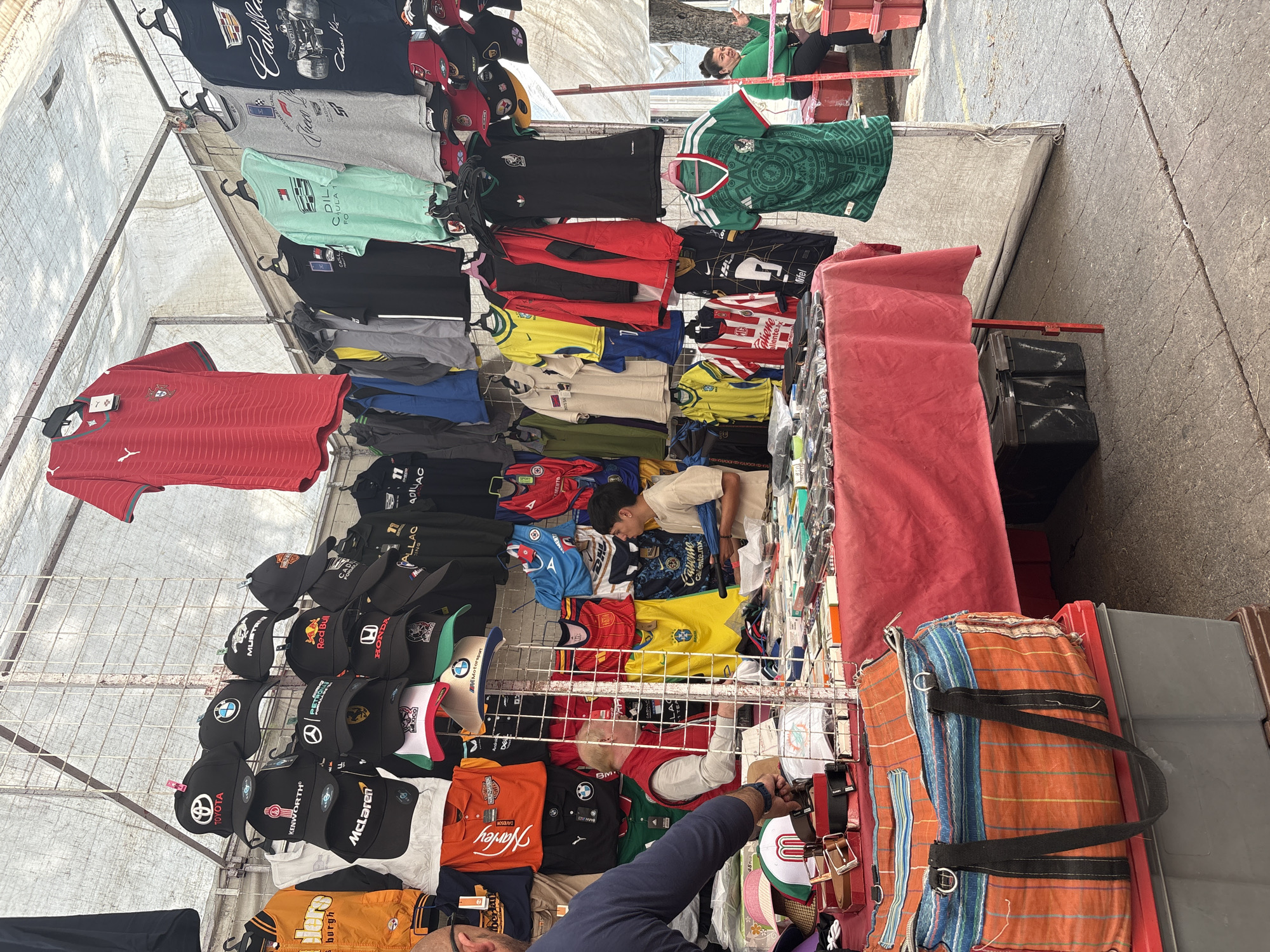 The image shows a street market in Mexico City, Mexico. The main focus is on a vendor's stall displaying a variety of merchandise, including shirts and hats. The shirts include soccer jerseys, racing team shirts, and various other designs. The hats are mostly racing team or brand-themed. A vendor is seen behind the display, seemingly tending to the arrangement of items. There are also two other people on the right of the stand, a person is bending over at the bottom, and another person is looking down. The stall is covered by a canopy and the environment appears to be a busy urban setting. A large patterned bag sits on a box on the right of the image.