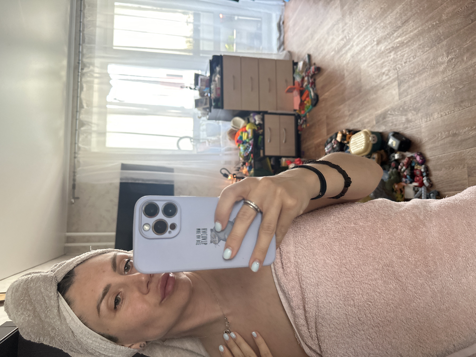 A woman with a towel wrapped around her head and body takes a mirror selfie in a room. She is wearing a light pink towel around her body and a beige towel on her head. The room appears to be a bedroom or living space, with wooden flooring. In the background, there are several drawers of a dresser, a television screen, and a window with sheer curtains. The floor is cluttered with various toys and objects, suggesting the presence of children. The overall lighting of the room is bright, likely from natural light entering through the window. The image does not provide enough information to determine the specific location within Novoaltaysk, Russia.