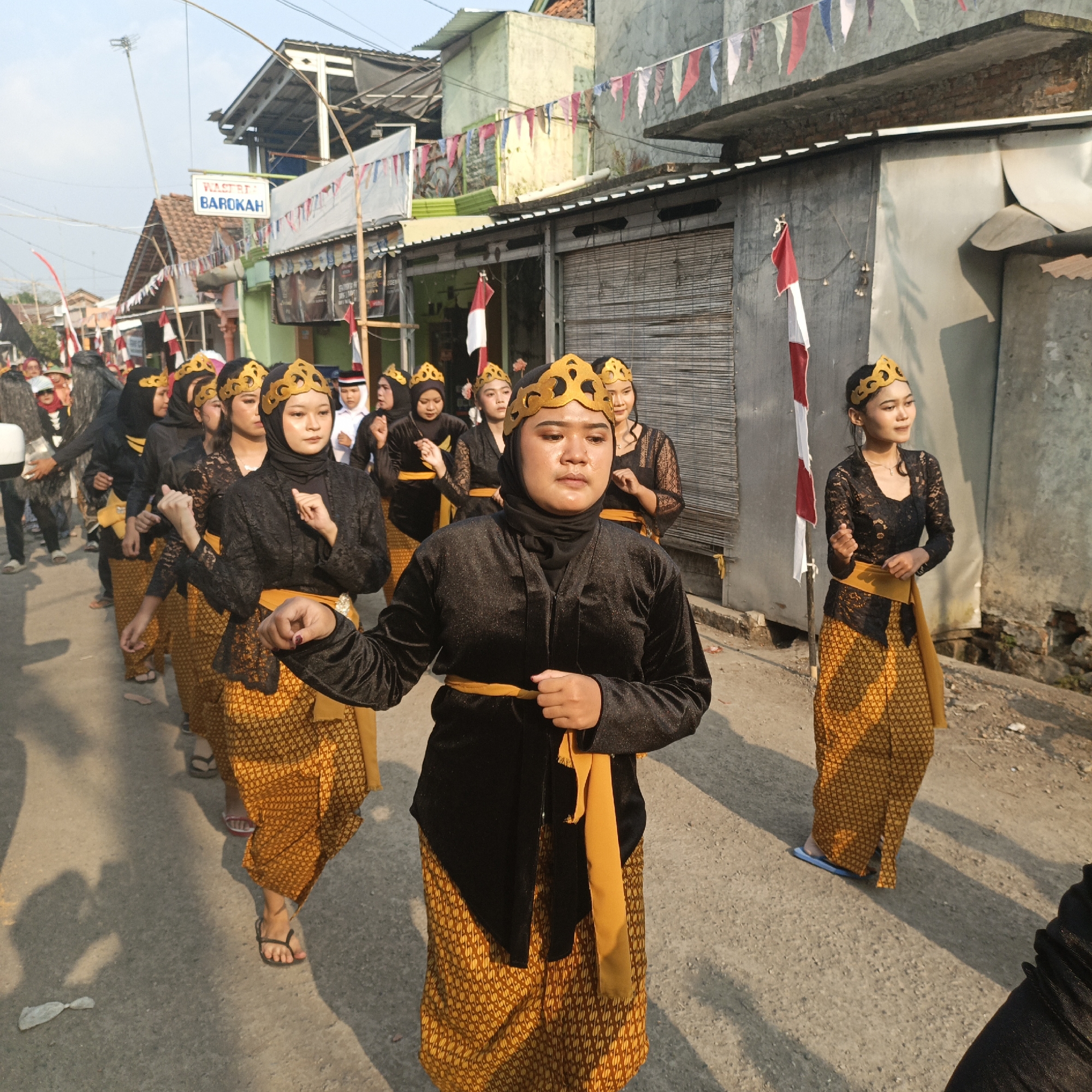 In Tanahbaya, Indonesia, a group of young women, adorned in traditional black attire with gold patterned skirts and golden headpieces, participate in a parade. The women are dressed in long-sleeved black tops and long skirts with a distinct gold and brown pattern. They are also wearing gold, crown-like headbands. Some are wearing black headscarves. A bright yellow sash is wrapped around the waist of each woman, with a portion hanging down. They appear to be moving in unison, possibly performing a dance or ceremonial walk.
The parade takes place on a paved street lined with buildings that have corrugated metal roofs and walls, some with open storefronts. Flags, including the Indonesian flag (red and white), are displayed along the street, adding to the festive or commemorative atmosphere. The sun is casting long shadows, indicating it is likely late afternoon or early morning. The environment appears to be a residential or commercial area within a town or city. A sign with white text on a blue background reading "WASTRA BAROKAH" is visible on a building in the background, suggesting a local business or establishment. The overall scene depicts a cultural event or celebration in a street setting in Tanahbaya.