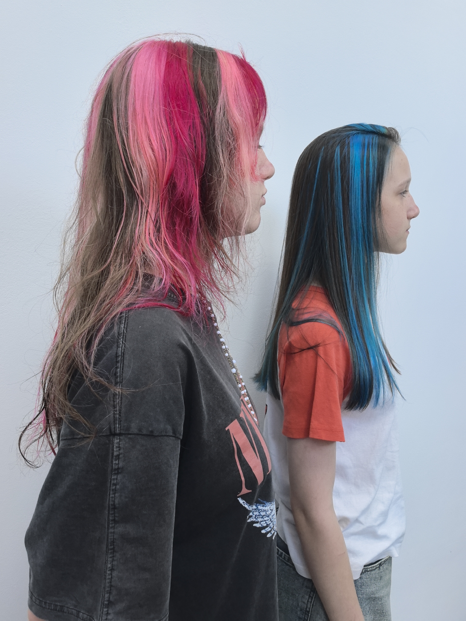 Two young people with brightly colored hair are standing in profile against a plain white background. The person in the foreground has long, wavy hair with streaks of hot pink and lighter pink. They are wearing a dark gray t-shirt with a graphic design on it. A beaded necklace is visible around their neck. The person in the background has dark hair with striking blue streaks. They are wearing an orange and white t-shirt and jeans. The image appears to have been taken indoors, with no specific environmental details to suggest a location in Chaikovsky, Russia.