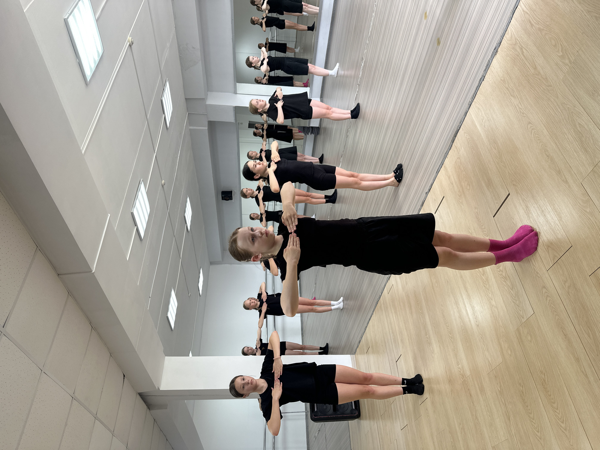 In a dance studio in Votkinsk, Russia, a group of young girls in matching black leotards and shorts are practicing a synchronized dance routine. They stand in two rows, facing the front, with their arms extended horizontally at chest level, palms down, in a balletic pose. The studio features a light-colored wooden floor, mirrored walls, and a white acoustic tile ceiling with fluorescent lighting. In the background, several other girls are visible, also engaged in the dance practice. A black speaker is positioned against the left wall. The overall impression is one of focused, disciplined movement and practice.