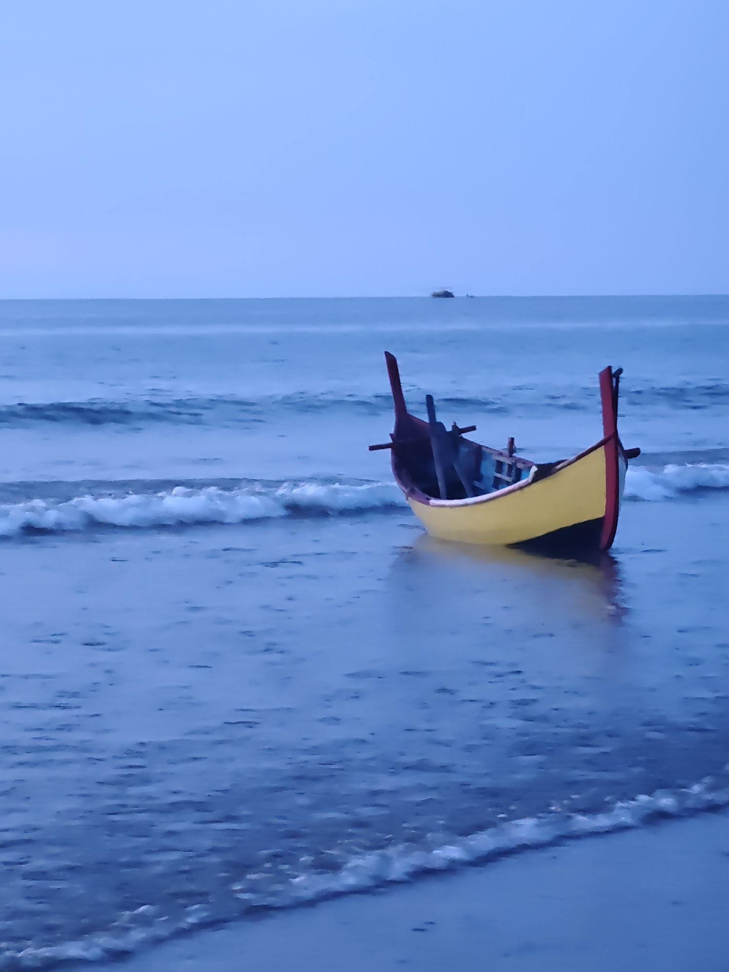 A yellow and red fishing boat rests on the shallow, wet sand at the edge of a calm ocean. The water reflects the overcast sky, which transitions from a pale blue at the horizon to a deeper indigo overhead. Small waves lap gently against the shore, creating a foamy white line where they meet the sand. The boat appears empty, with its dark interior contrasting with its bright exterior. The scene is serene and quiet, suggesting either dawn or dusk. There are no people visible in the image. This scene is located in Curehtunong, Indonesia.