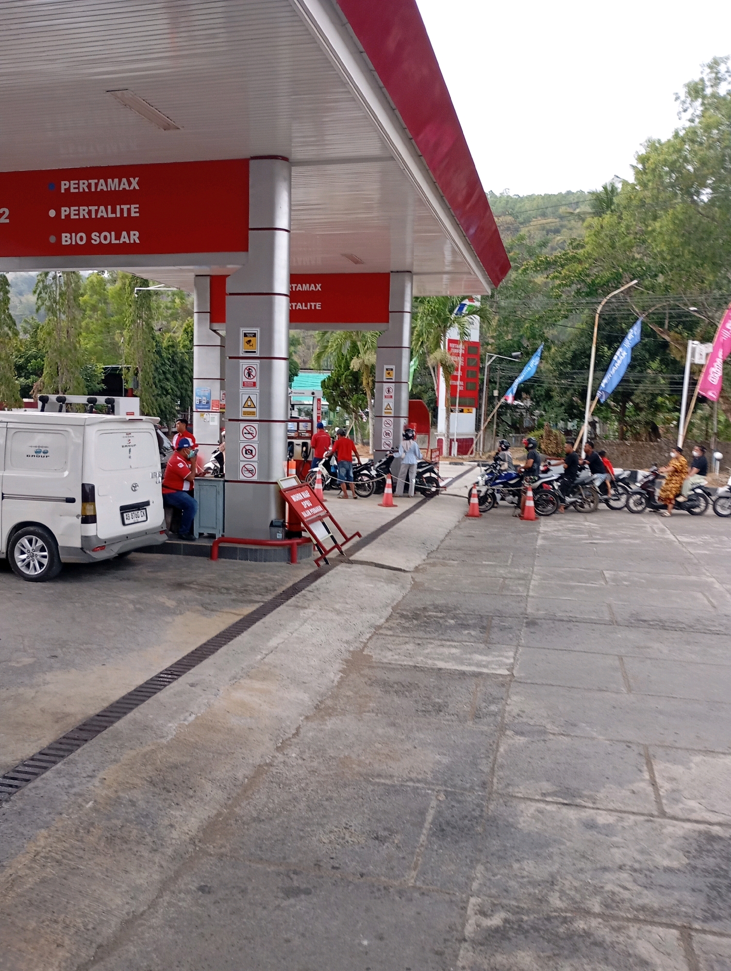 This image depicts a petrol station in Menadi, Indonesia. The station features a red and white canopy with signs indicating "PERTAMAX," "PERTALITE," and "BIO SOLAR." Several people are present, with some operating motorcycles at the pumps. A white van is parked to the left, and various safety signs and flags are visible around the station. The ground is paved with concrete and stone tiles, and a drainage grate runs along the edge of the paved area. The background shows lush green trees and a hilly landscape, suggesting a location outside a densely urbanized area. The overall scene captures a typical transaction at a gas station, with vehicles and individuals engaging in the refueling process.
