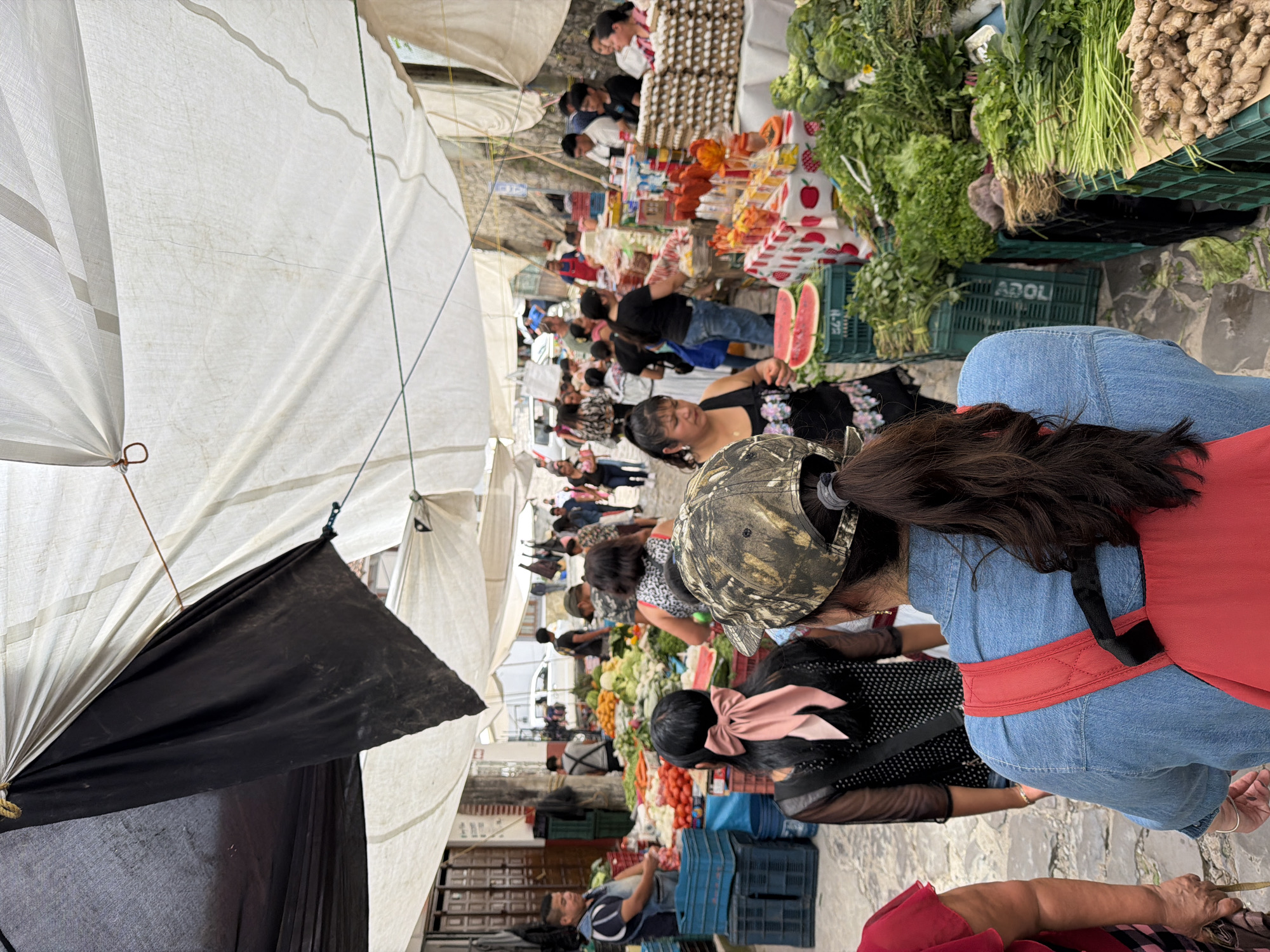 The image depicts a bustling market in Cuetzalan, Mexico. People are browsing and purchasing goods from vendors under large, white canopies. There are various stalls displaying produce like fruits, vegetables, and eggs. Customers are wearing casual clothing, and some are carrying bags or backpacks. The environment appears crowded with people interacting at the stalls. The pavement is visible. A person is seen wearing a denim jacket with a red backpack. Another person has a camouflage hat, and yet another wears a bow in their hair.