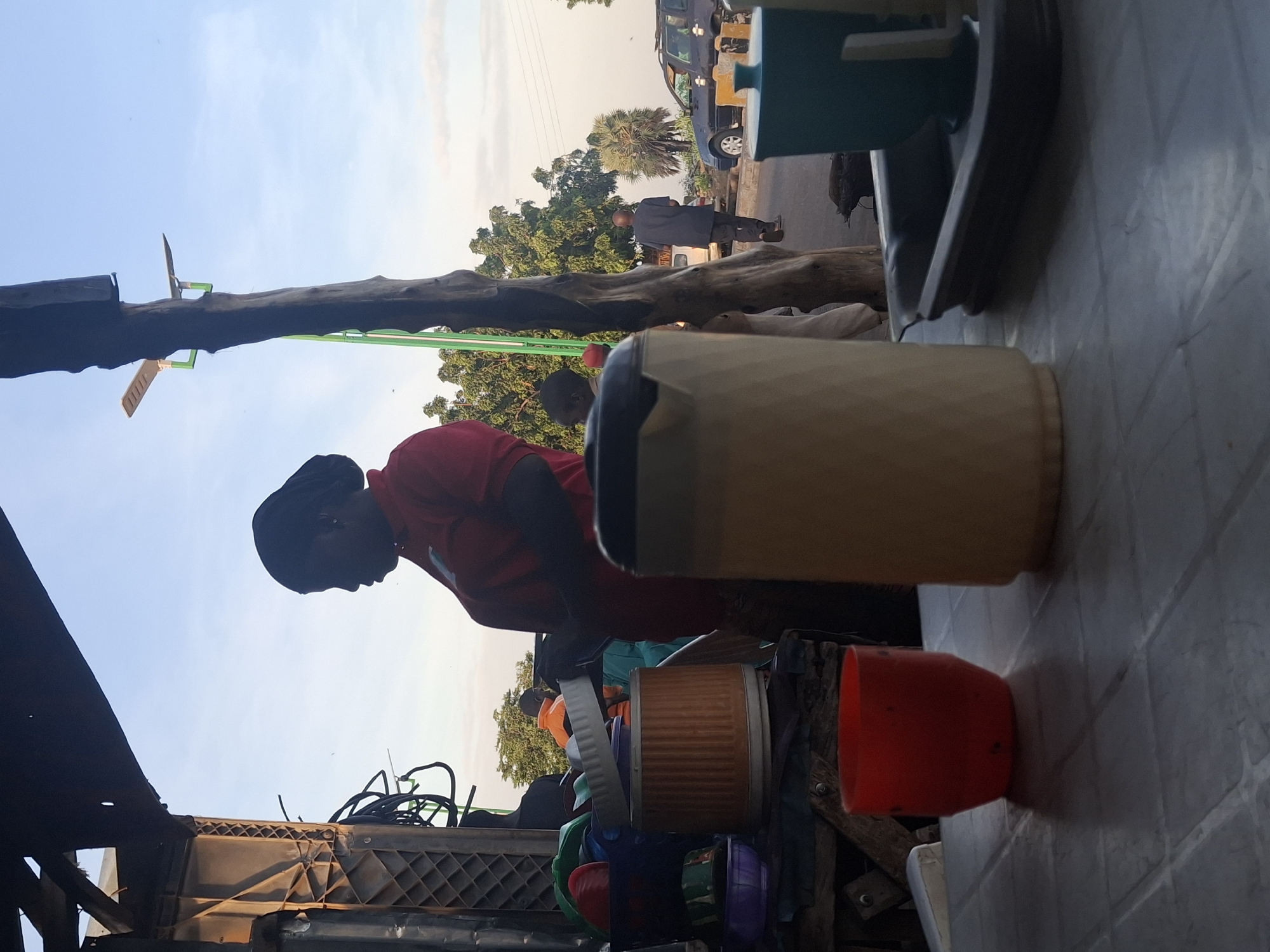 In Gombe, Nigeria, at dusk, a woman wearing a red shirt and headwrap is seen from the side. She is standing behind a table laden with various plastic containers. In front of her are a red cup, a large beige pitcher, and a dark green container with a light lid on a grey tray. The setting appears to be an outdoor stall or shop, with a rough wooden structure overhead and a pathway or road visible in the background. A man in a dark shirt and trousers walks away from the camera on this road, and a parked dark blue car is also visible. The sky is clear with a faint glow of sunset.