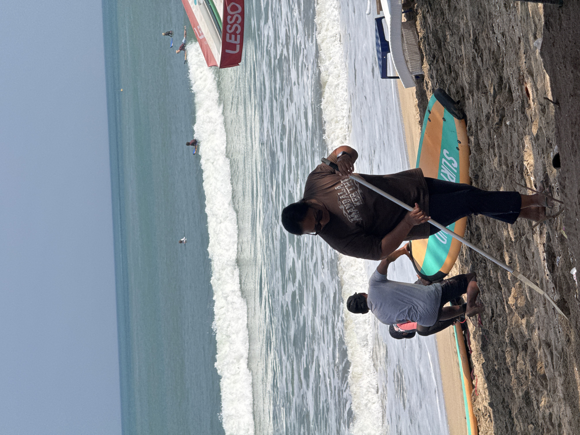 A group of individuals are on the sandy shore of Kuta Beach, Bali, Indonesia, near the ocean on a sunny day. The ocean has moderate waves, and several surfers are visible in the water. In the foreground, a person in a brown t-shirt and dark pants is using a long pole to clear debris from the sand. To their left, another person in a grey t-shirt and dark shorts is kneeling next to a surfboard. Another individual, partially visible, is also near the surfboard. Two surfboards are lying on the sand. Beach chairs and a red and white umbrella are situated further back on the beach. The sky is clear and blue.