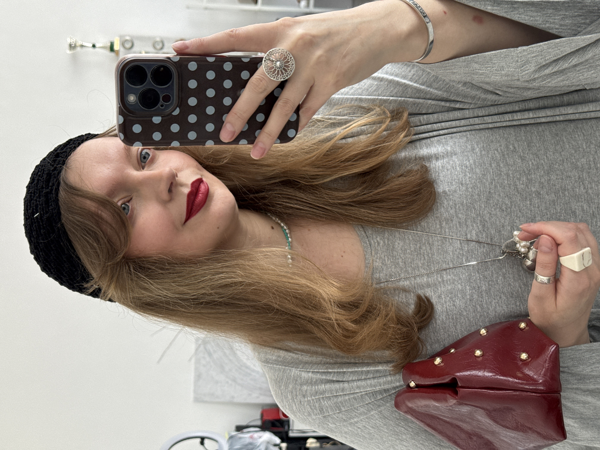 A young woman with fair skin, blue eyes, and shoulder-length blonde hair, wearing a black crocheted beanie and red lipstick, is taking a selfie. She is holding a smartphone in her left hand, which is adorned with a large, circular ring. The phone has a brown case with light blue polka dots. She is wearing a light gray long-sleeved top. Her right hand is near a small, red, studded clutch purse. She is also wearing a silver bracelet and silver rings. A silver necklace is visible around her neck. The background appears to be a white wall with a blurry reflection of a decorative item on the upper left. A portion of a shelf with various items on it is visible at the bottom of the frame. There is no indication of the location being Izhevsk, Russia, in the image.