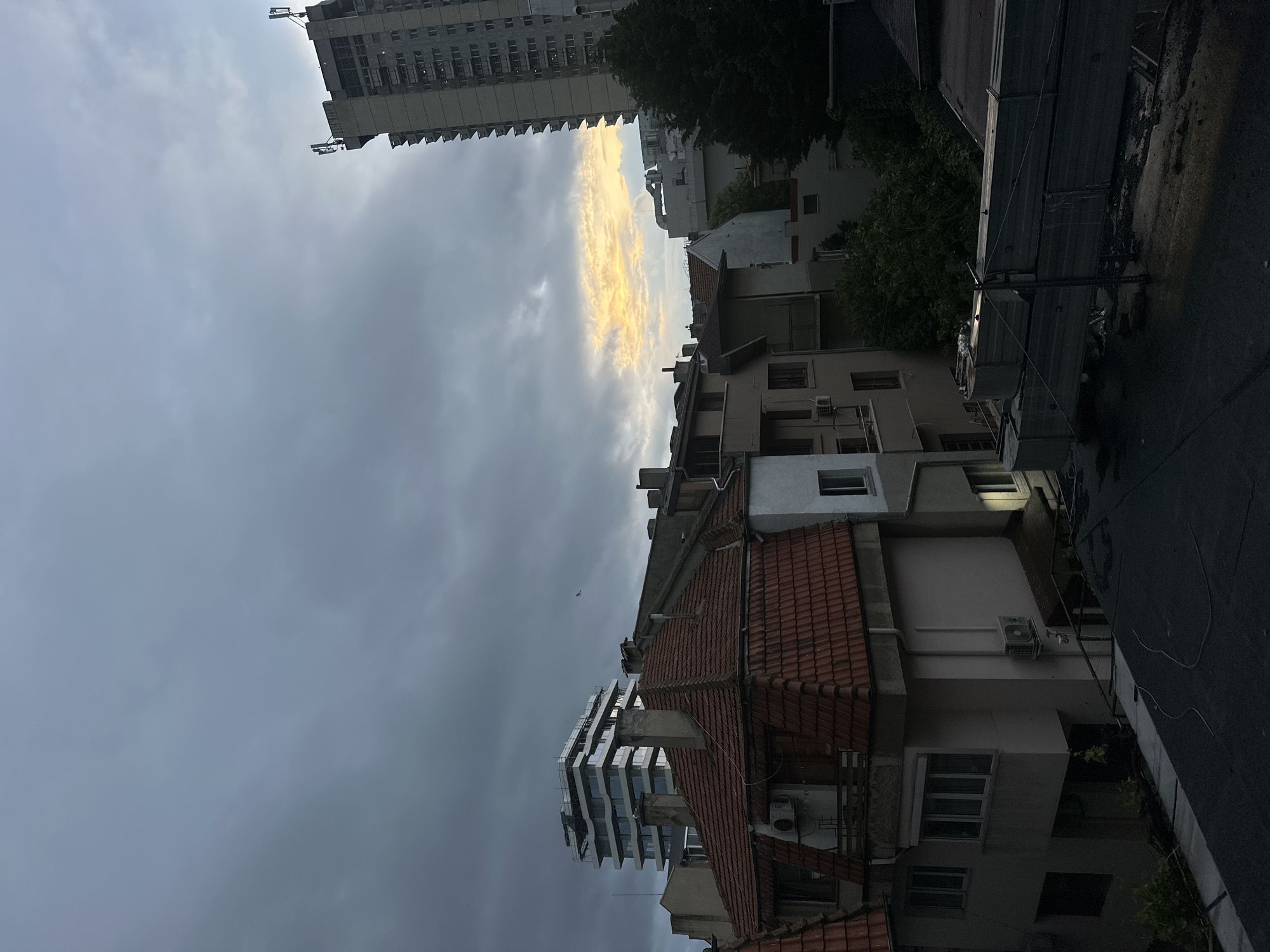 The image captures an urban scene in Varna, Bulgaria. In the foreground, buildings with red-tiled roofs and aged facades dominate. These older structures are interspersed with more modern apartment buildings. The sky is overcast with heavy, grey clouds, although a sliver of golden light breaks through the horizon, suggesting either sunrise or sunset. The narrow street below is dimly lit and appears to be wet. A utility pole with wires is visible. The overall impression is that of a city undergoing a transition, with older and newer structures coexisting under a dramatic sky.