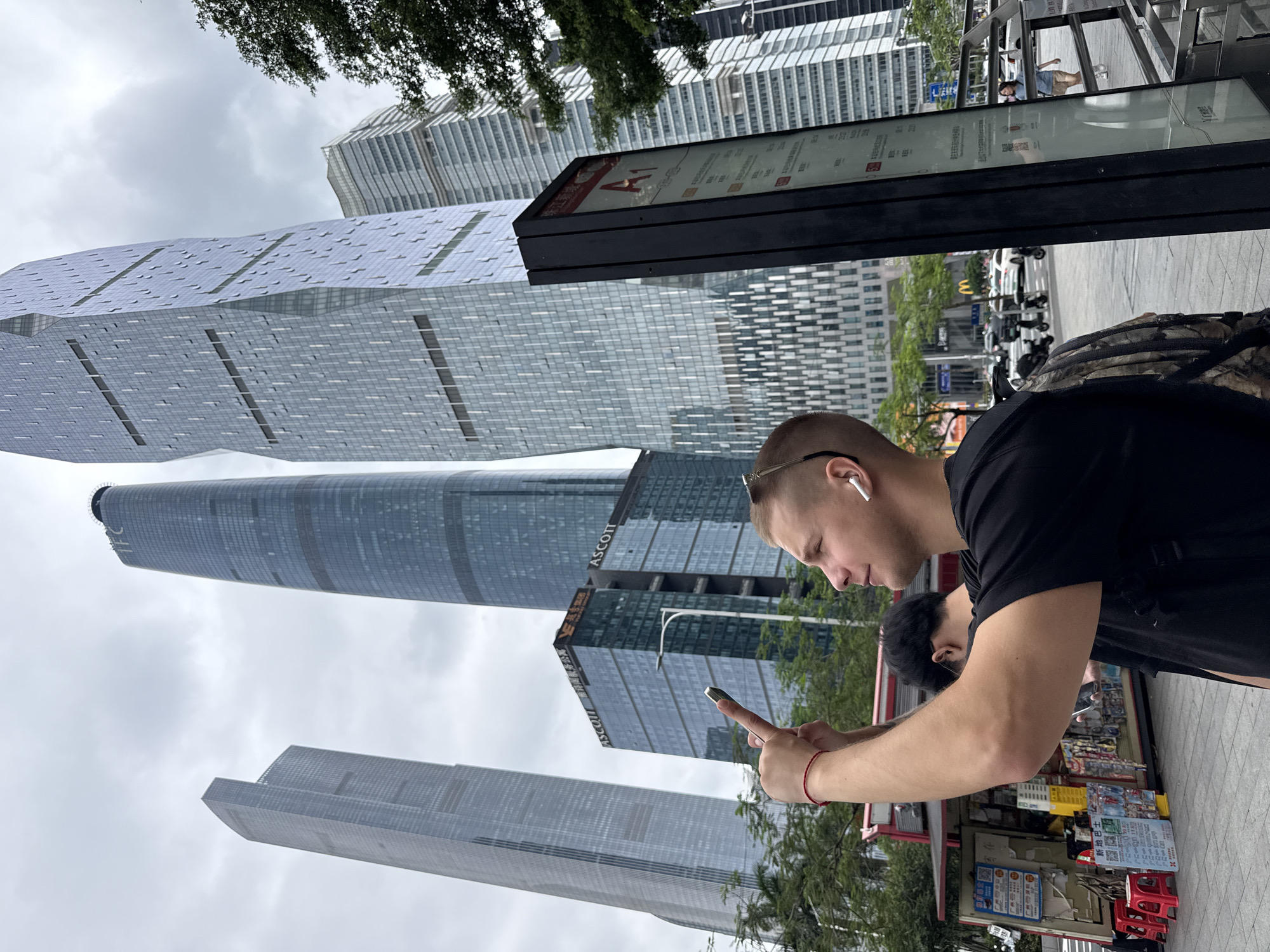 In the Tianhe District of China, under an overcast sky, a young Caucasian man with short blonde hair and a backpack is taking a photo with his phone. He is wearing a black t-shirt and white wireless earbuds. His right arm is extended upwards, holding the phone. In the background, several modern skyscrapers dominate the cityscape. To the left of the man, a newsstand is visible. A signpost with a red "A1" is positioned to the right of the man. The street in front of the skyscrapers is busy with traffic, including cars and scooters.
