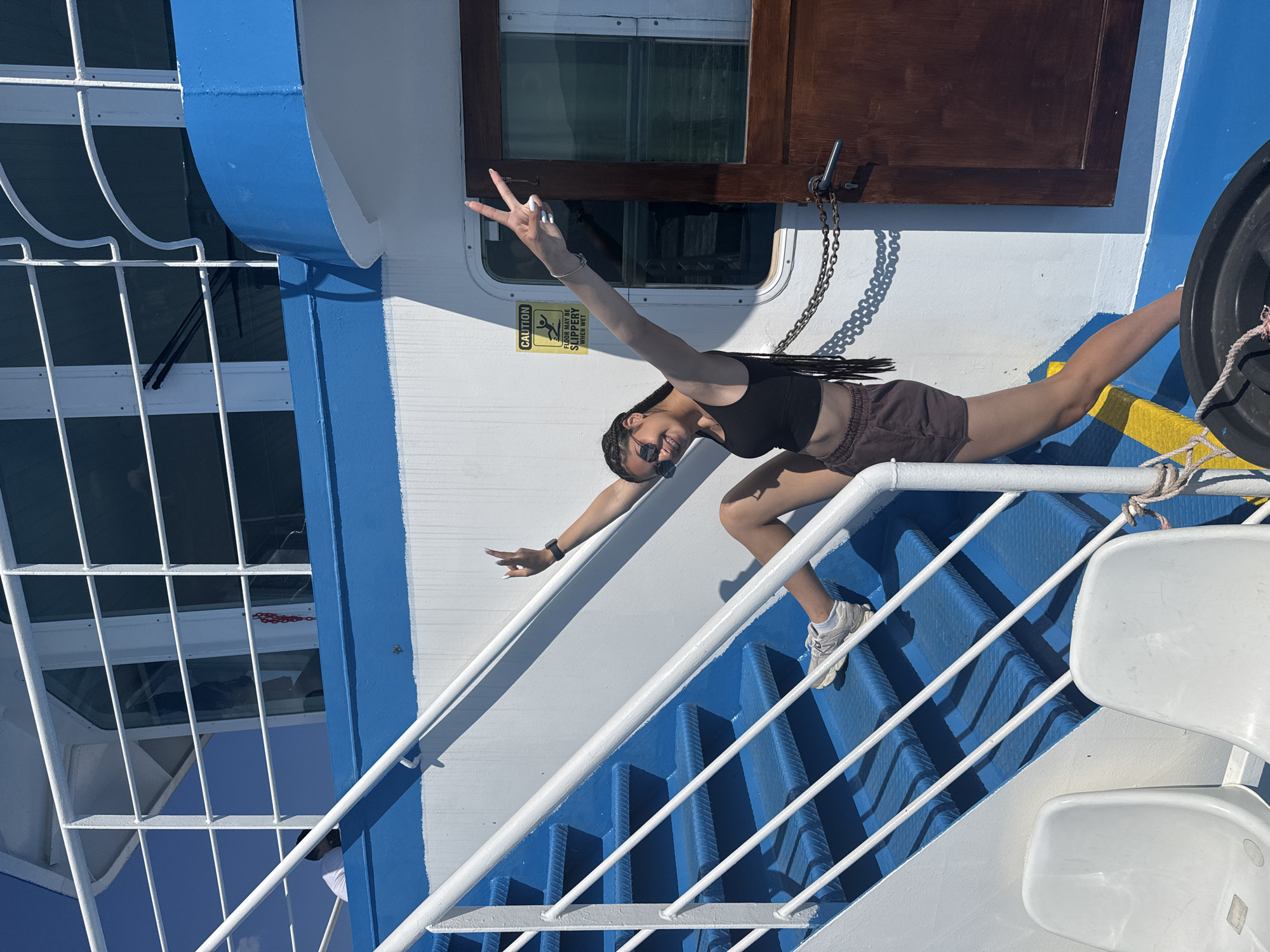 A young woman with dark braided hair is posing on the blue stairs of a white and blue ferry in Greece. She is wearing a black crop top, brown shorts, and white sneakers. She is smiling and holding up two peace signs with her hands. The sun is shining, and the sky is blue. There are white railings on the stairs. A "CAUTION: FLOOR MAY BE SLIPPERY WHEN WET" sign is visible on the wall of the ferry.