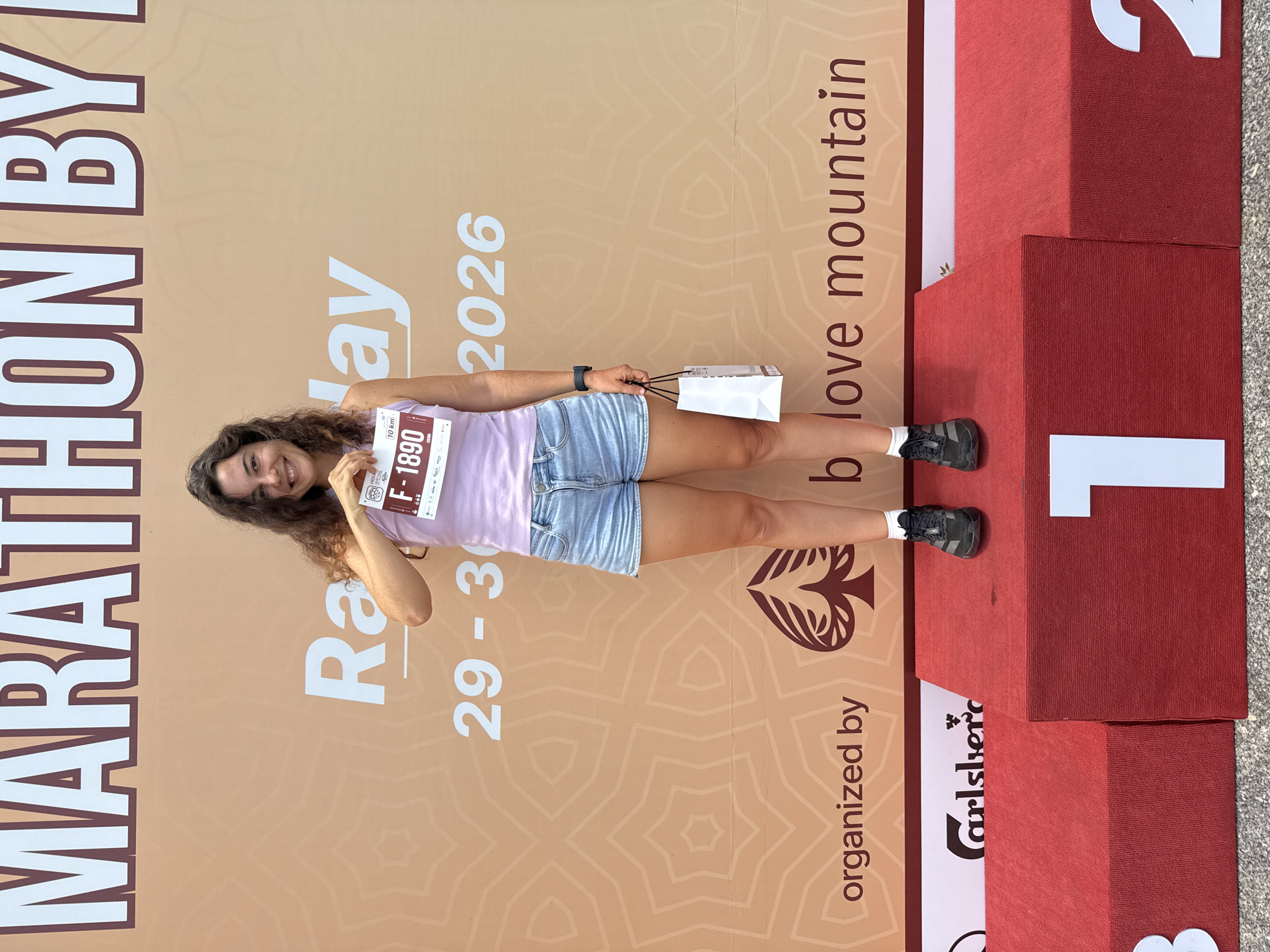 A woman stands on a podium in front of a banner in Hoi An, Vietnam. She is wearing a light purple tank top, denim shorts, and athletic shoes. She is holding a race bib with the number F-1890. The banner behind her reads "MARATHON BY" at the top and "Race Day" in larger font below. The date "29 - 30" and the year "2026" are also visible on the banner. The bottom of the banner has logos and text that says "organized by b love mountain". A red podium with the numbers 1, 2, and 3 on it is in the foreground. The woman is smiling and looking at the camera.