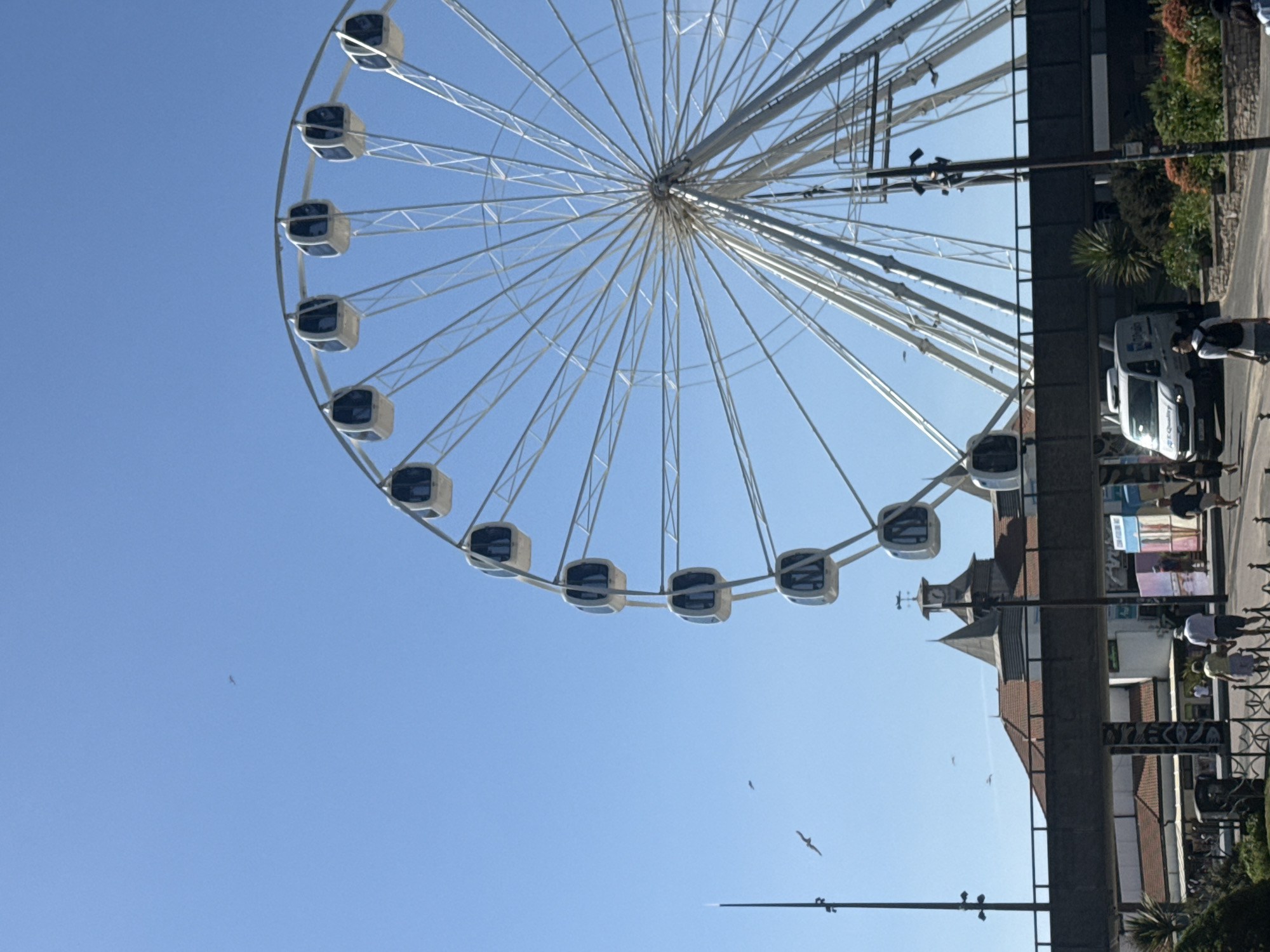 The media file depicts a Ferris wheel against a clear blue sky, suggesting a sunny day. The Ferris wheel itself is a prominent object, with its spokes extending outwards and several gondolas visible. To the right of the Ferris wheel, there is a building with a brick facade and a pointed roof. A street runs alongside the building, with people walking and a white van parked. The setting appears to be a coastal town, possibly a seaside resort, given the presence of a Ferris wheel, which are often found in such locations. The image has been rotated, with the Ferris wheel positioned on the left and the buildings and street on the right. Some birds are visible in the sky. The overall impression is one of a pleasant, possibly tourist-oriented location on a bright day.
